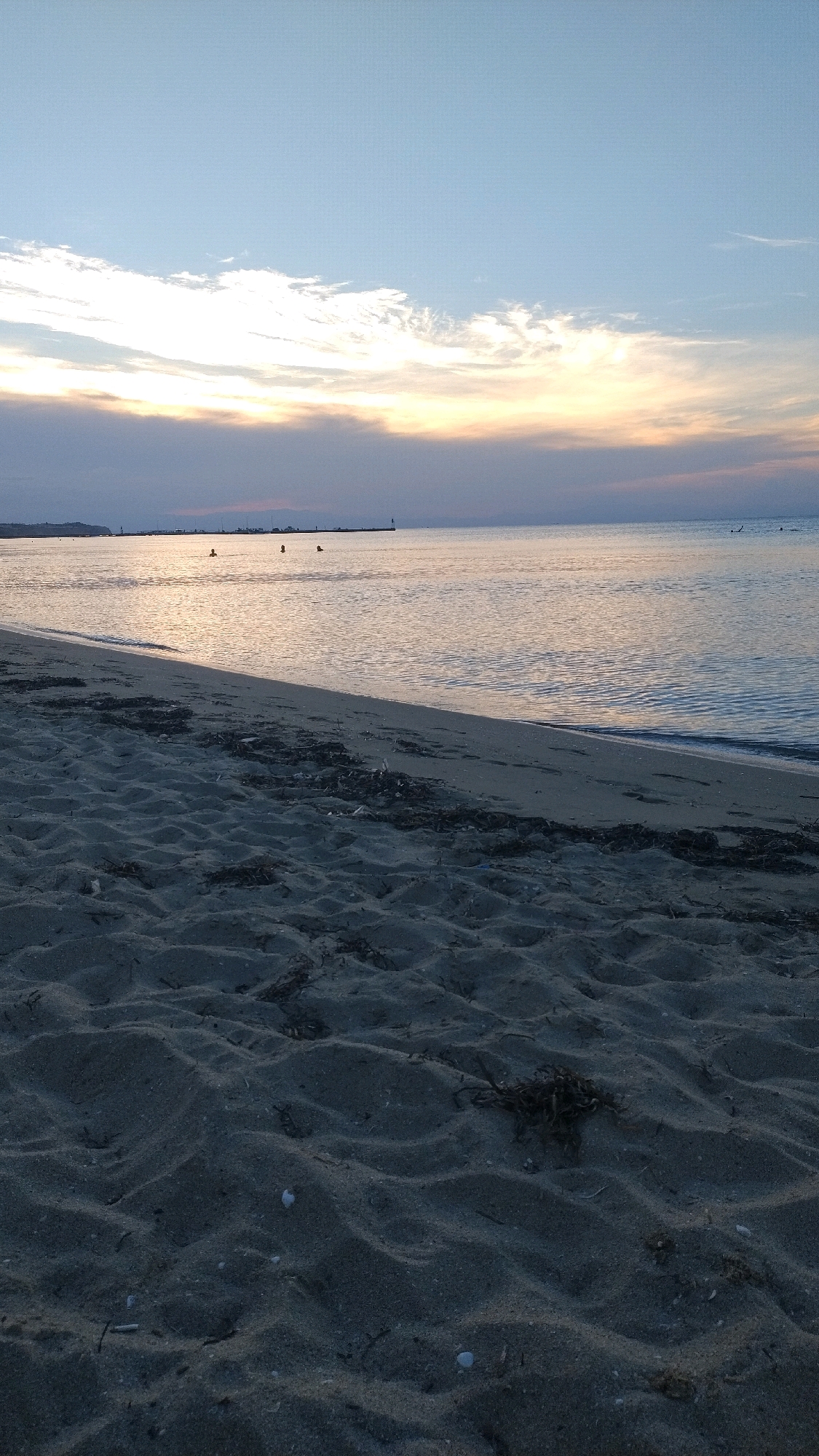 The image depicts a serene beach scene at dusk in Neoi Epivates, Greece. The foreground is dominated by textured sand, showing the subtle contours of dunes and scattered dried seaweed. The waterline gently laps at the sandy shore. In the mid-ground, several individuals are visible in the calm, reflective water, appearing to be swimming or wading. A distant pier extends into the sea, marked by a small lighthouse. The horizon is defined by a band of soft clouds, illuminated by the fading sunlight, which casts a warm, golden glow on the water's surface. The sky above transitions from pale yellow near the horizon to a clear, soft blue.