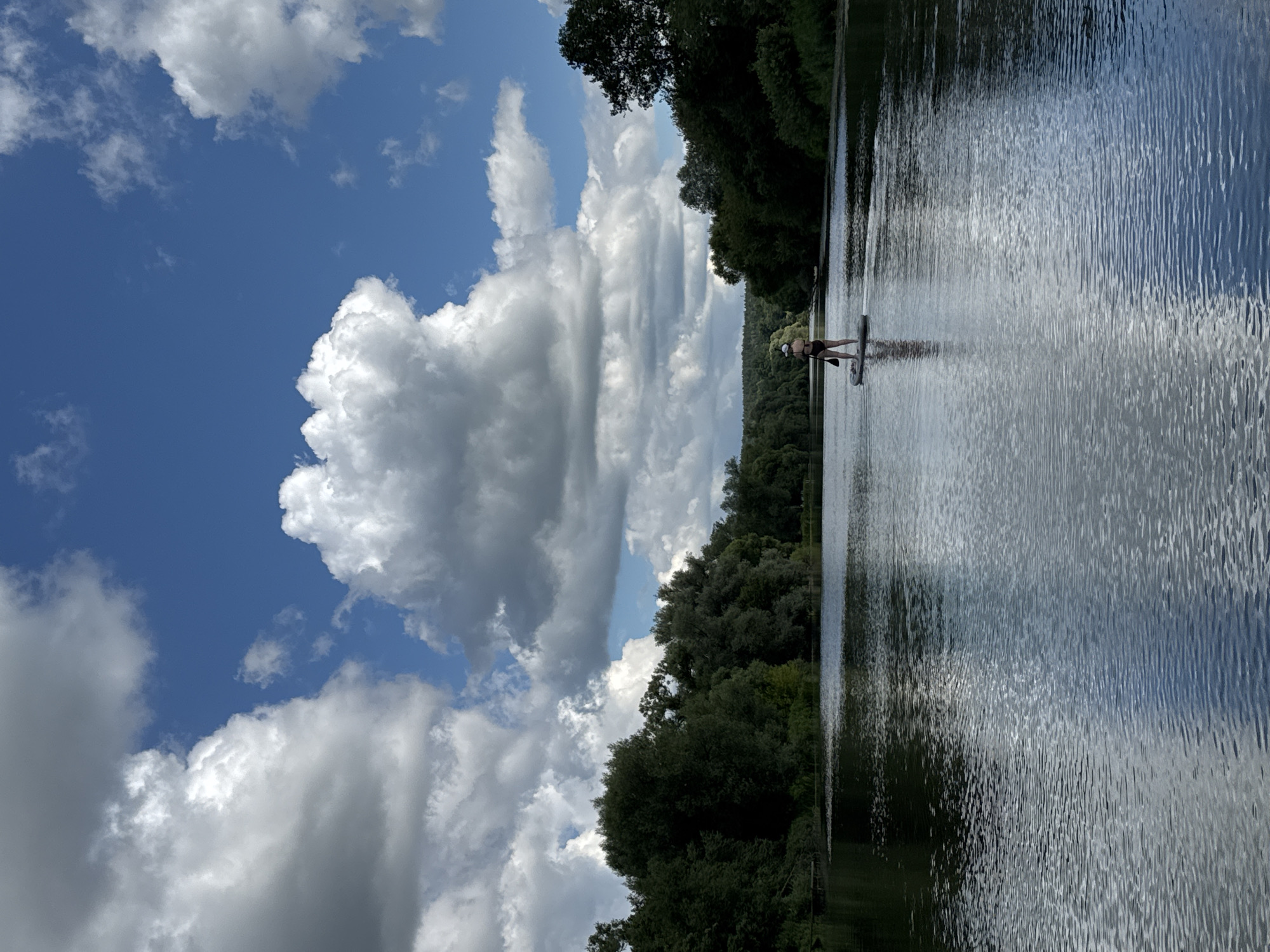 A lone individual is paddleboarding on a calm river, surrounded by lush green trees on both banks. The sky is a vibrant blue, adorned with large, fluffy white clouds. The water reflects the sky and trees, creating a serene and picturesque scene. The individual is standing on a paddleboard, holding a paddle, and appears to be paddling forward. The time of day is daytime, indicated by the bright sunlight. The location appears to be a natural body of water, possibly a river or lake, with dense vegetation along the shorelines. The weather is clear and sunny, with some clouds adding visual interest to the sky.