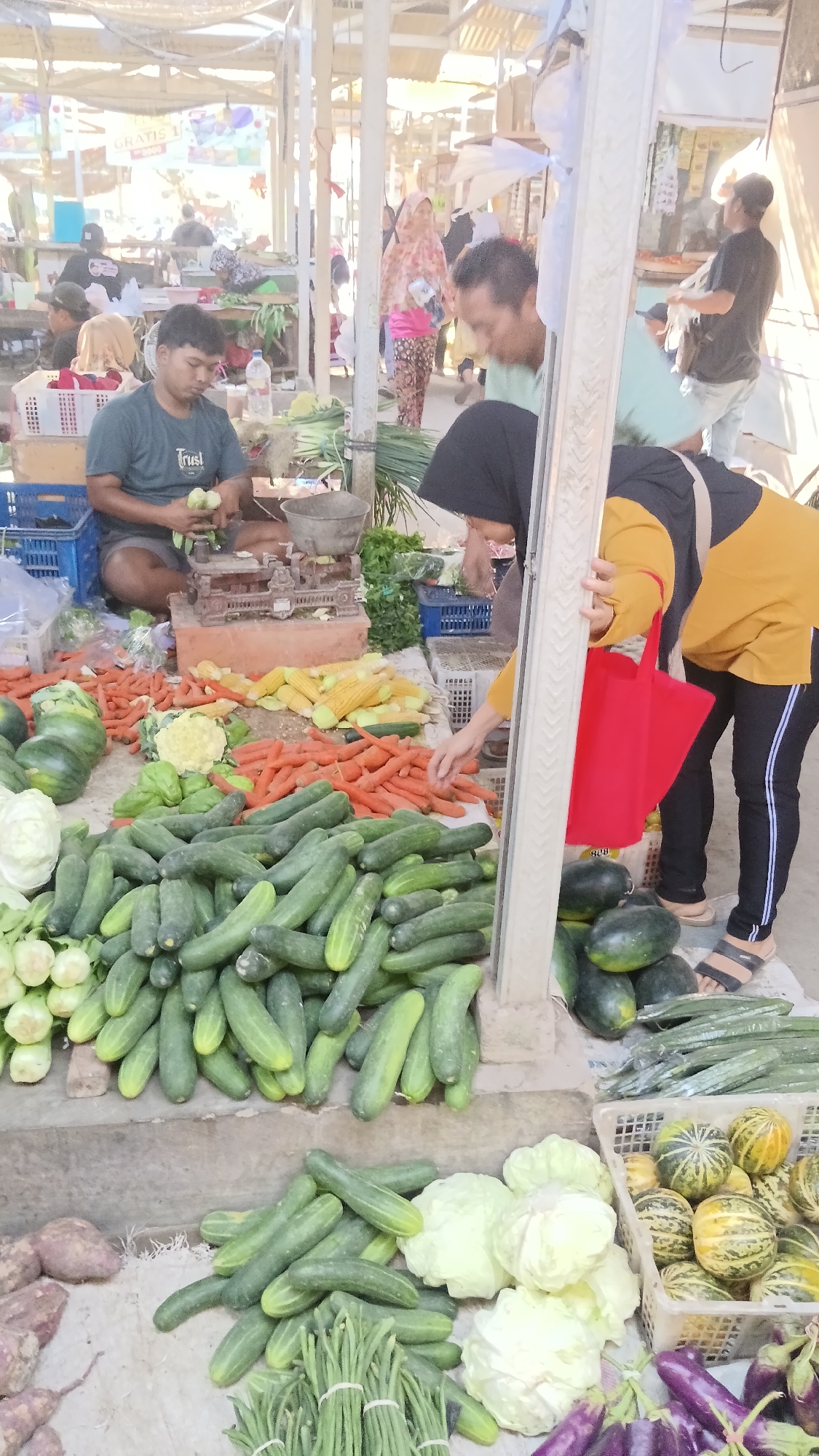 At an outdoor market in Dukuh, Indonesia, a man in a green t-shirt is weighing vegetables on a scale. He is surrounded by piles of fresh produce, including cucumbers, carrots, corn, cabbage, and what appear to be sweet potatoes. A woman in a yellow shirt and black pants is reaching for vegetables, holding a red bag. In the background, other people are browsing the market stalls under a covered structure. The scene is bustling with activity, indicating a typical day at a local Indonesian market.