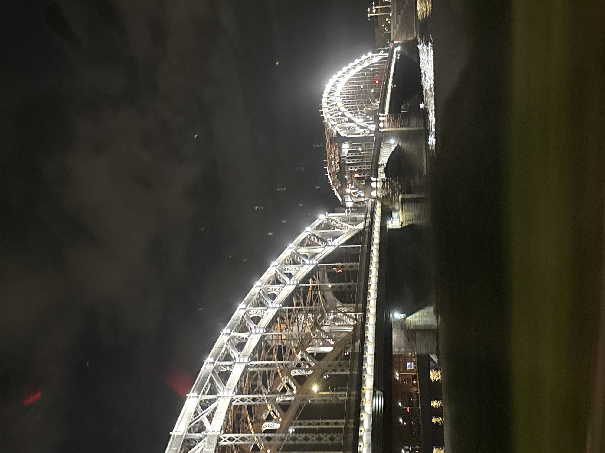 This is a night photograph of the Bolsheokhtinsky Bridge in Saint Petersburg, Russia. The bridge, also known as the Peter the Great Bridge, is a prominent landmark spanning the Neva River. The image captures the illuminated steel arches of the bridge against a dark, cloudy sky. The lights on the bridge are bright white, highlighting its intricate metalwork. The Neva River below reflects the city lights, creating shimmering patterns on the water's surface. In the foreground, there is a dark, out-of-focus area that appears to be a grassy embankment. The overall scene conveys a sense of urban grandeur and nocturnal beauty.