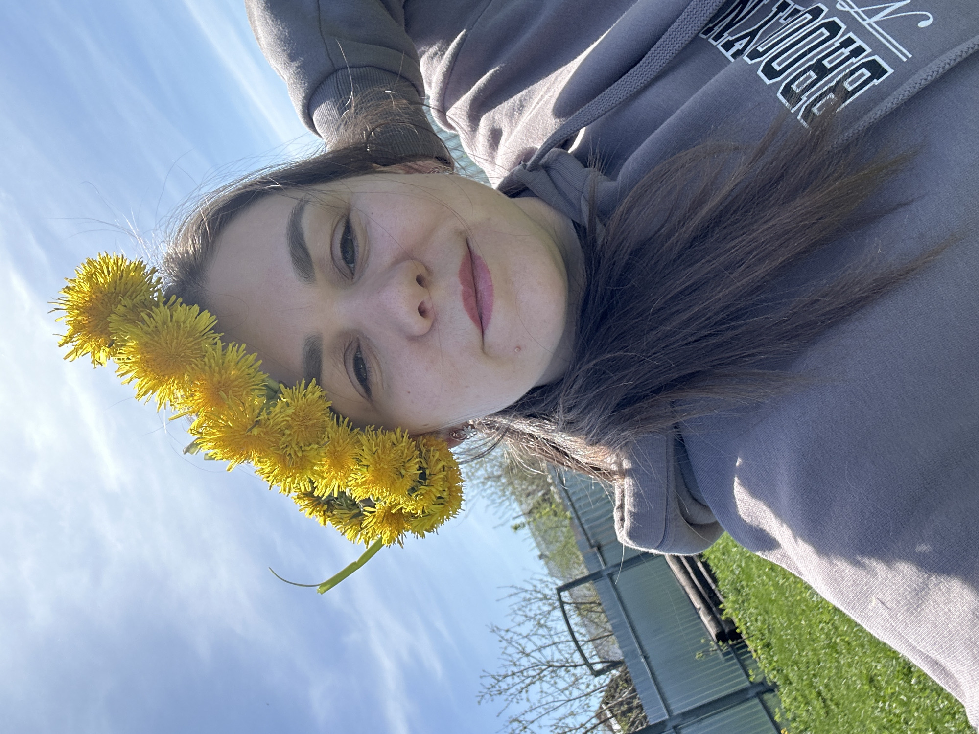 A woman wearing a gray hoodie with "BROOKLYN" printed in white letters, and a crown of yellow dandelions on her head, is positioned at an angle in an outdoor setting. The sky, featuring some clouds, is visible in the background. The location is identifiable as Chaikovsky, Russia due to the presence of Russian flora and fauna as well as the style of the surrounding architecture. Fencing and greenery are partially visible in the background as well.