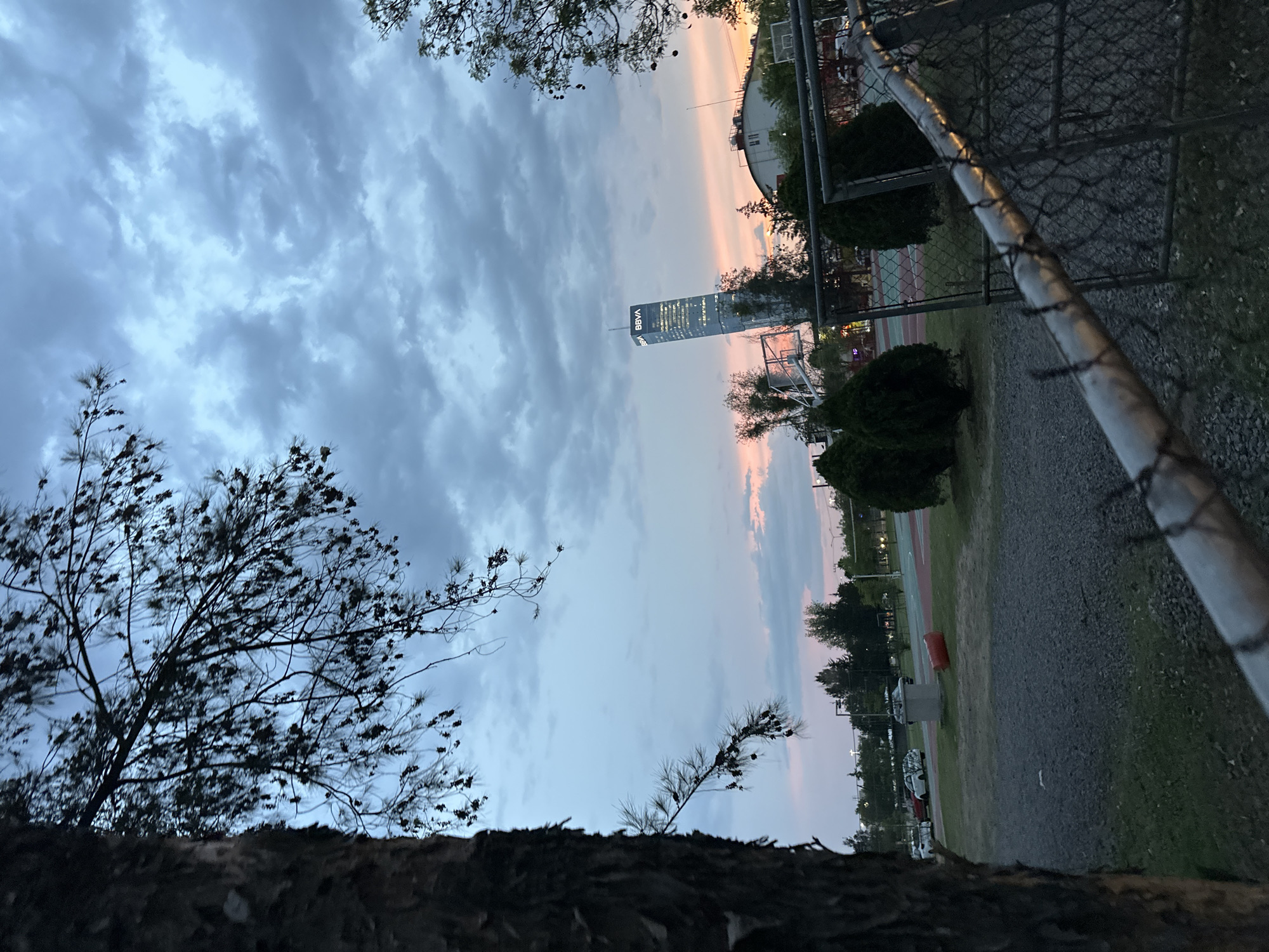 This media file captures an outdoor urban landscape scene at dusk or sunset in San Bernardino Tlaxcalancingo, Mexico. The sky is a prominent feature, displaying a soft gradient of blues, grays, and delicate pink and orange hues indicative of twilight. Numerous clouds, varying in shape and density, are scattered across the sky.
The central subject is a tall, modern skyscraper, illuminated with internal lights, standing prominently against the colorful evening sky. The white text "BBVA" is clearly visible near the top of the building.
In the foreground and mid-ground, a mix of natural and man-made elements is present. To the left, silhouetted trees with dark, slender branches and needle-like leaves frame the view. On the right, a sturdy chain-link fence, topped with barbed wire, diagonally traverses the frame, indicating a boundary. Beyond the fence, the ground consists of a gravelly path leading to a grassy area. Elements of a recreational space are partially visible, including what appears to be part of a basketball court and a hoop. Bushes and additional trees are scattered throughout the background behind the fence.
No people or explicit activities are depicted, contributing to a calm and serene atmosphere. The artificial lights of the BBVA building contrast with the fading natural light, marking the transition into evening. The scene offers a view of urban development integrated with a natural or park-like setting.
