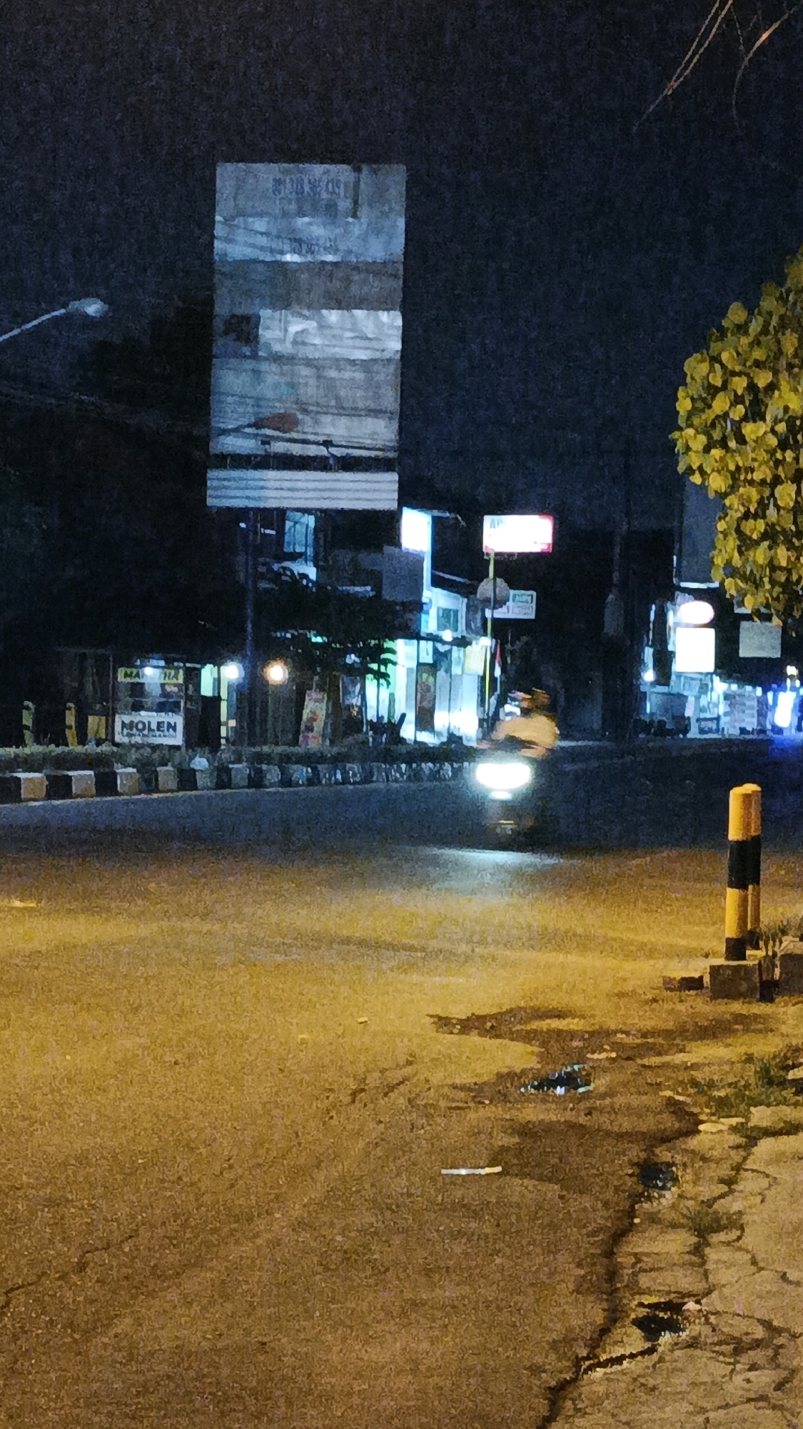 This is a nighttime, eye-level, medium shot of a street scene in Surakarta, Indonesia. The road surface is asphalt, with some cracked sections and puddles reflecting light. On the right side of the road, there is a yellow and black striped pole, likely a traffic marker, and a concrete curb. Across the street, there are several buildings with illuminated signs, suggesting commercial activity. One sign clearly reads "MOLEN" in large white letters on a blue background. A street lamp is visible on the left side of the image. A vehicle, its headlights illuminated, is moving on the street. The overall atmosphere is dark, with artificial lighting from the buildings and vehicles creating contrast.