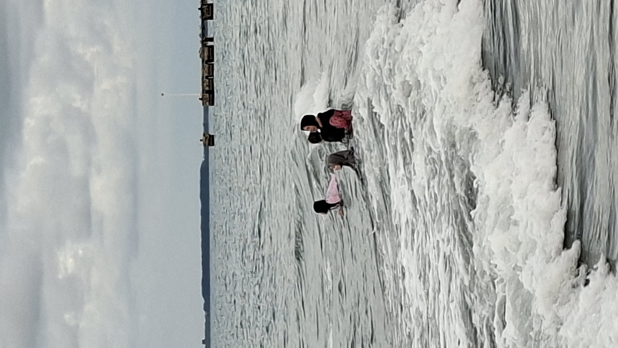 The image depicts a group of four individuals, appearing to be children or young women, in the water at a beach in Lhokseumawe, Indonesia. They are partially submerged, with waves breaking around them. Three of the individuals are wearing hijabs, and one is in a pink top, while another is in a dark top with pink accents. They are positioned in the water, with some of them lying on their stomachs. In the background, there is a body of water with a distant shoreline and a pier-like structure. The sky is overcast.