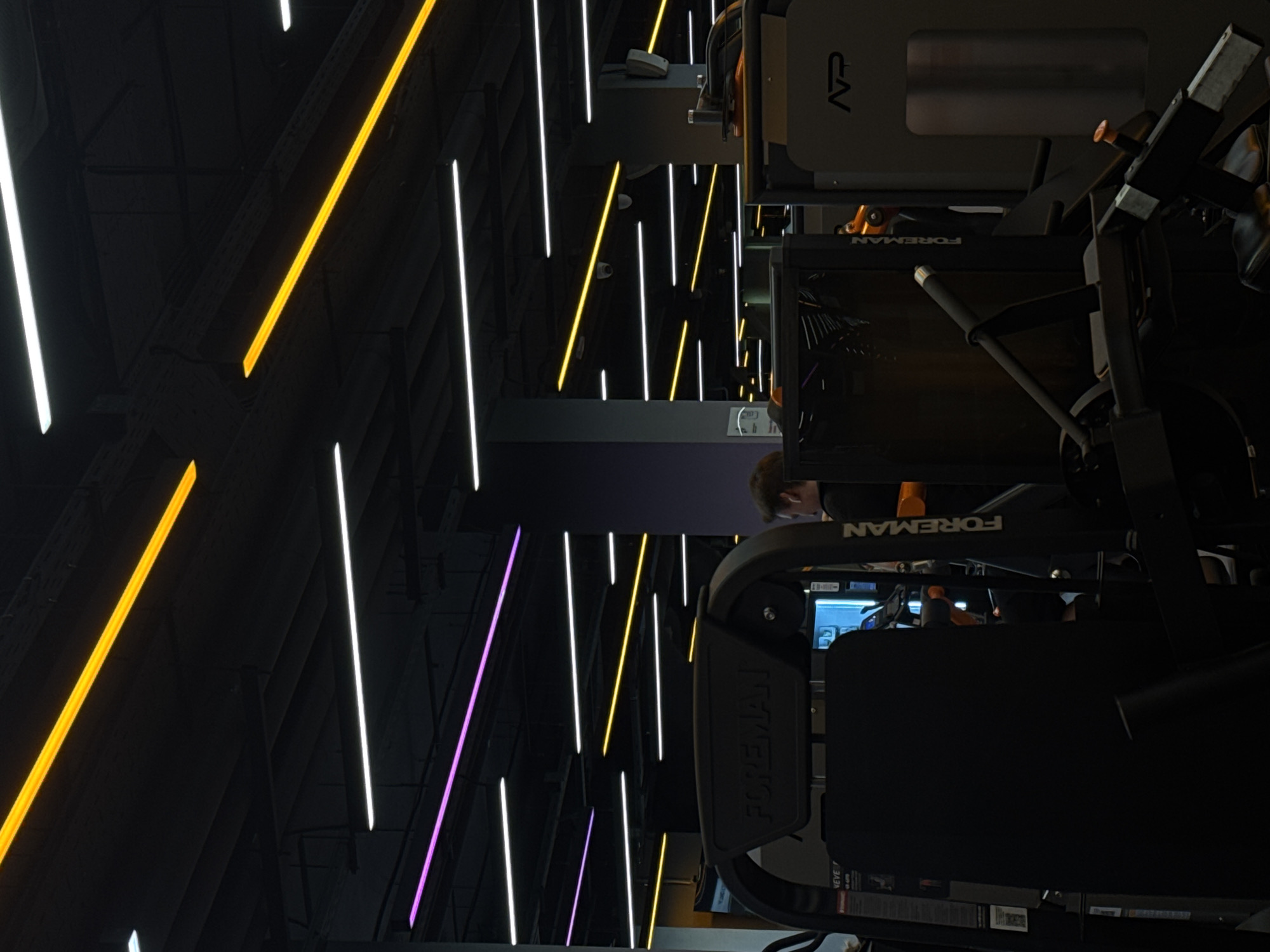 The image depicts the interior of a gym, likely in a commercial setting. Several pieces of workout equipment, including machines labeled "FOREMAN," are visible in the foreground and midground. A person with short brown hair is partially visible in the background, near what appears to be a dark purple section of the wall.
The environment features a dark ceiling with parallel lines of colored light: yellow, white, and purple. These lights run vertically across the image and create a distinct visual effect, suggesting a modern or stylized design. The background is mostly dark, with the colored lights providing the main illumination.
The location is undetermined, but it's not in Saint Petersburg, Russia. The image does not contain any information which could tie it to Saint Petersburg or Russia in general.