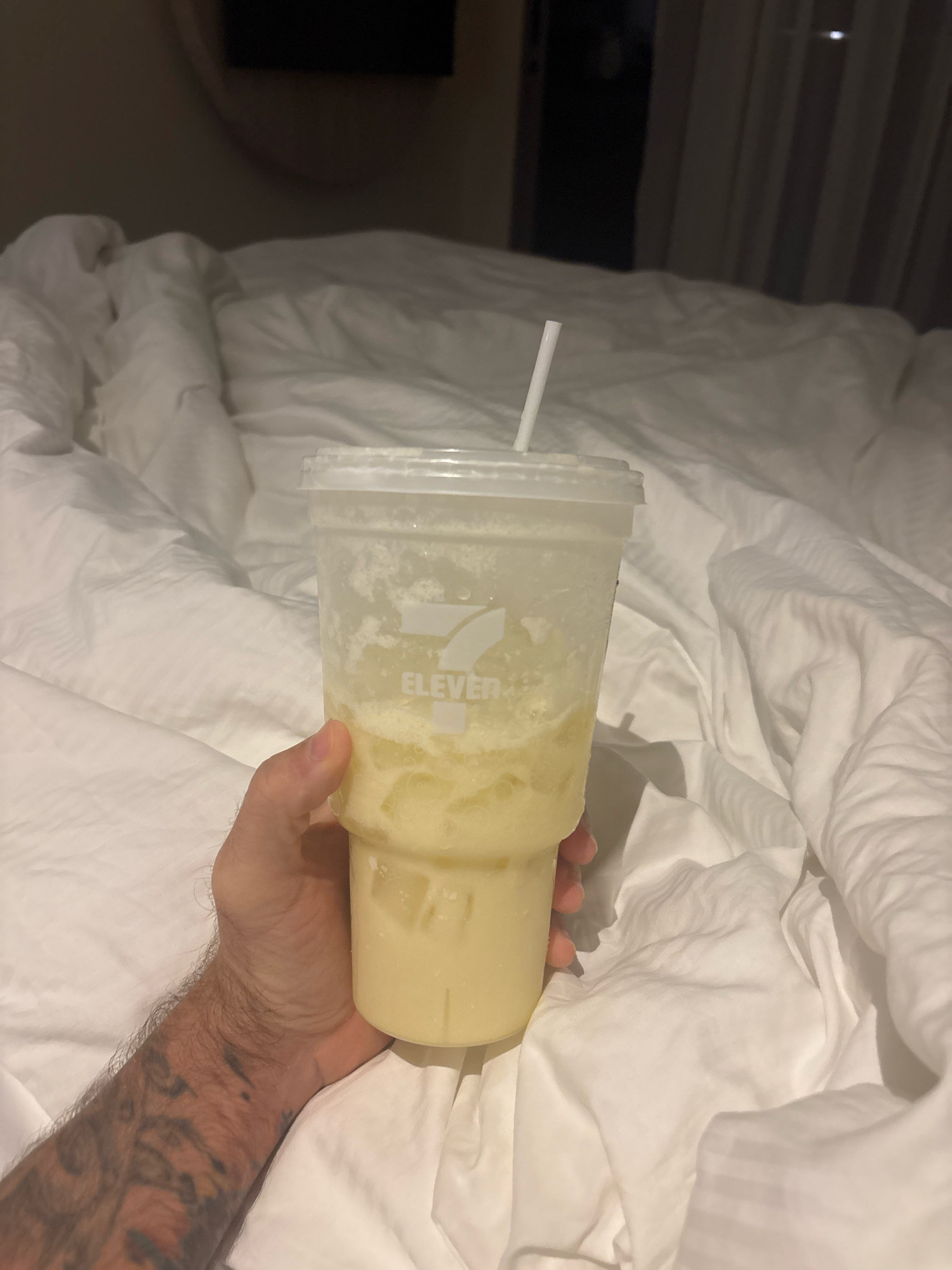 An overhead, medium shot captures a hand holding a clear plastic cup filled with a pale yellow, slushy beverage. The cup, bearing the "7-ELEVEN" logo, has a white lid and a white straw protruding from it. The beverage contains ice cubes and appears frothy. The hand holding the cup is tanned and has visible tattoos on the forearm. The background consists of neatly folded white bedding, suggesting the scene is likely in a hotel room. The overall lighting is dim, with some ambient light reflecting off the sheets and the cup. The location is specified as Karon, Thailand, implying the bed and bedding are in a lodging establishment within that area.