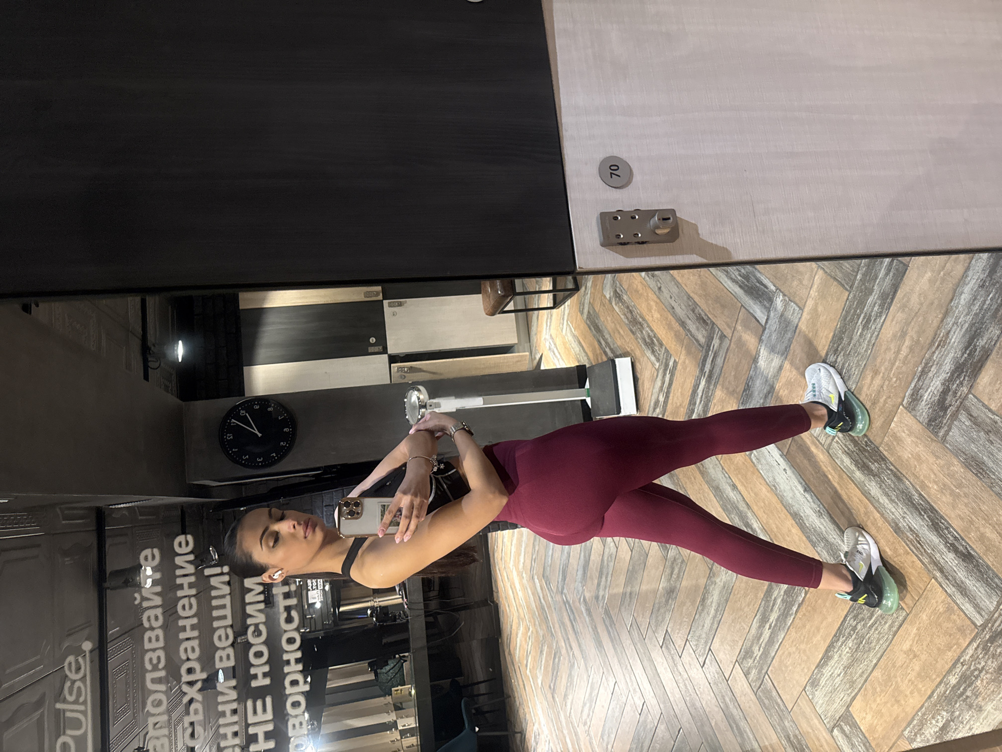 A young woman is taking a selfie in what appears to be a locker room or gym area. She is wearing a black top and burgundy leggings. She is holding a smartphone in her left hand and seems to be posing for the mirror reflection. The background includes lockers and a wall with text in Cyrillic alphabet ("Ползвайте", "съхранение", "енни вещи!", "НЕ НОСИМ", "Оговорност!"), which can be translated from Bulgarian to English as: "Use", "Storage", "Valuable things!", "We are not responsible!". A clock on the wall displays the time. The flooring is a light-colored herringbone pattern. It is implied that this scene is in Sofia, Bulgaria.