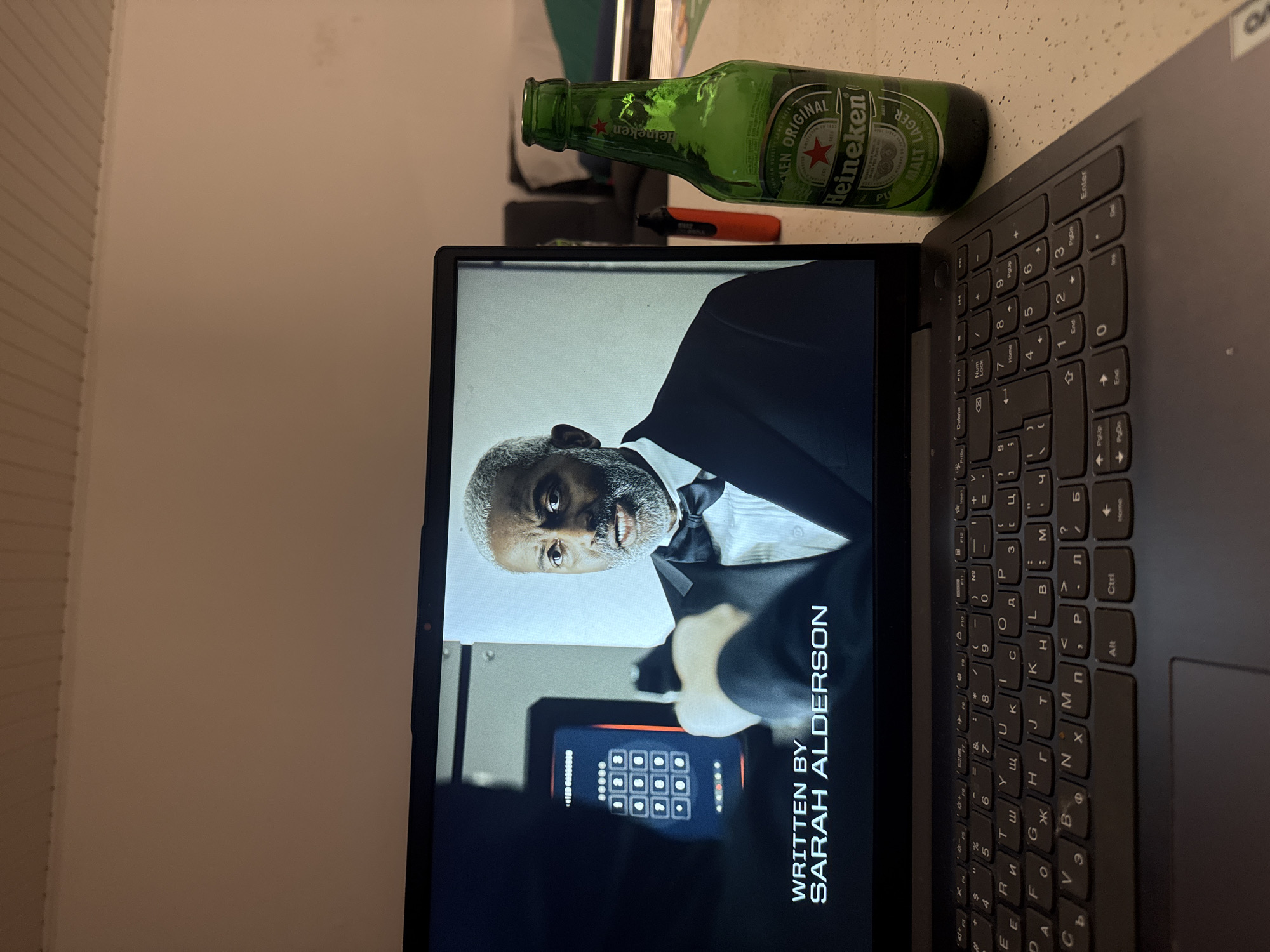 The media file presents an indoor scene, likely in Hattersheim am Main, Germany, captured from a rotated perspective. In the foreground, a dark-colored laptop is open on a speckled, light-colored surface. Its keyboard features both Latin and Cyrillic characters. The laptop's screen displays a frame from what appears to be a film or television show. The central figure on the screen is a man with grey hair and a beard, wearing a black tuxedo with a white shirt and bow tie, looking directly forward with a serious expression. Below him, part of a digital keypad, possibly for a safe or entry system, is visible. Overlaid text on the screen clearly reads "WRITTEN BY SARAH ALDERSON."
Adjacent to the laptop, a green glass bottle of Heineken Original beer is prominently displayed. The bottle is mostly full, with its label clearly legible. The background shows a plain, light beige wall, suggesting a simple domestic or office setting. A small orange object is subtly visible behind the laptop screen in the distance. The overall scene suggests an individual relaxing and consuming media, possibly during the evening or late hours, indicated by the indoor lighting.