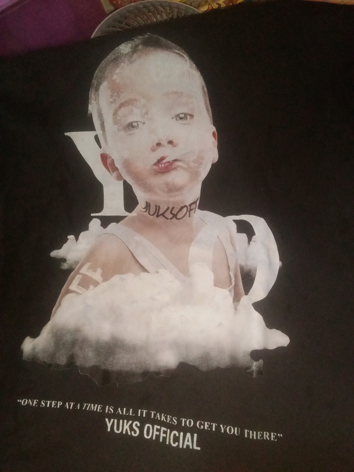 The image displays a black t-shirt with a graphic design. The design features a baby's face, appearing as if covered in a white powder or paint, with its mouth puckered. The baby is wearing a light-colored top and has a white "Y" shape behind its head. Text is visible on the t-shirt, reading "YUKSOFF" around the baby's neck, and at the bottom, there is text that says "ONE STEP AT A TIME IS ALL IT TAKES TO GET YOU THERE" and "YUKS OFFICIAL". The image is a close-up shot of the t-shirt, likely taken indoors. There is no information in the image that indicates a location in the city of Karawang, Indonesia.