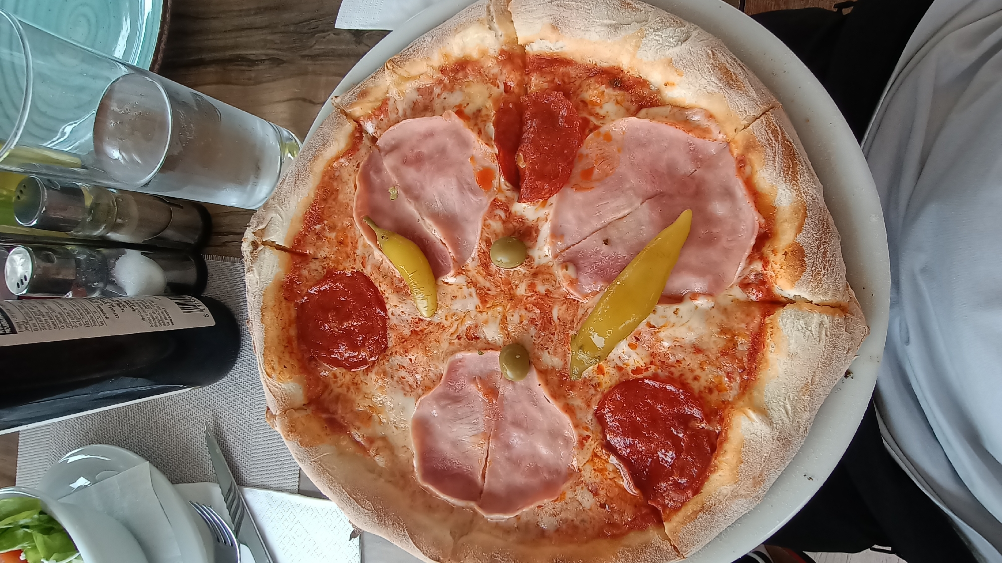 The scene depicts an overhead view of a dining table, identified as being in Umag, Croatia. The primary subject is a large, circular pizza on a white ceramic plate, positioned centrally. The pizza features a thick, lightly browned crust, a tomato sauce base, and melted white cheese. Toppings include four slices of light pink ham, four slices of red pepperoni or salami, two whole green olives, and two elongated yellow pickled peppers. The pizza is pre-cut into at least eight slices.
Surrounding the pizza, on a light-colored woven placemat, are various dining items. To the upper left, a white bowl containing a green salad is partially visible, alongside a small white saucer and a fork and knife set resting on a white paper napkin. In the upper-center, a dark glass bottle with a white label displaying an 8-digit barcode and text, partially obscured, stands upright. Adjacent to the bottle are two small glass shakers with metal tops, one containing white granular salt and the other a darker granular substance, likely pepper. To the right of the shakers, a tall, clear glass filled with water and ice stands, exhibiting condensation. A section of a light blue-green plate is visible in the upper right background. A small, crumpled white paper napkin rests on the right edge of the pizza plate. At the bottom of the frame, the lap and lower torso of a person, wearing a light-colored t-shirt and dark pants, are visible, suggesting a first-person perspective of a diner at the table. The table surface beneath the placemat appears to be made of wood.