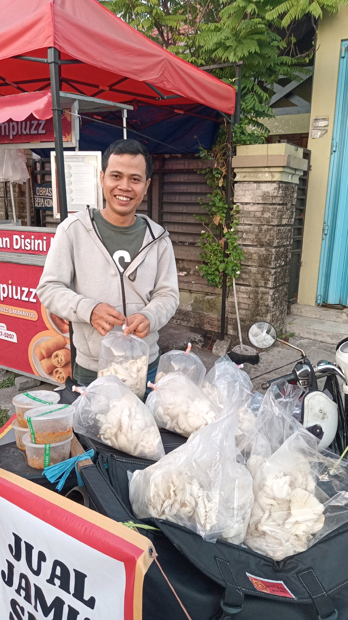 A medium-shot photograph captures a smiling Indonesian man standing behind a food stall in the city of Randuagung, Indonesia. The man, wearing a light grey hooded jacket over a green t-shirt, is holding two plastic bags filled with food items. He is positioned in front of a red and black food stall canopy that has signage that reads "mpıuzz". To his right, a motorcycle is partially visible. The stall is laden with multiple clear plastic bags containing what appear to be dried mushrooms, as well as several small plastic containers of sauce. In the background, a brick building with a light blue door and a small tree with green leaves are visible. The man's hands are near the opening of the plastic bags he is holding, suggesting he is in the process of packaging or preparing food for sale. The lighting suggests it is daytime, possibly late afternoon, with diffused natural light.