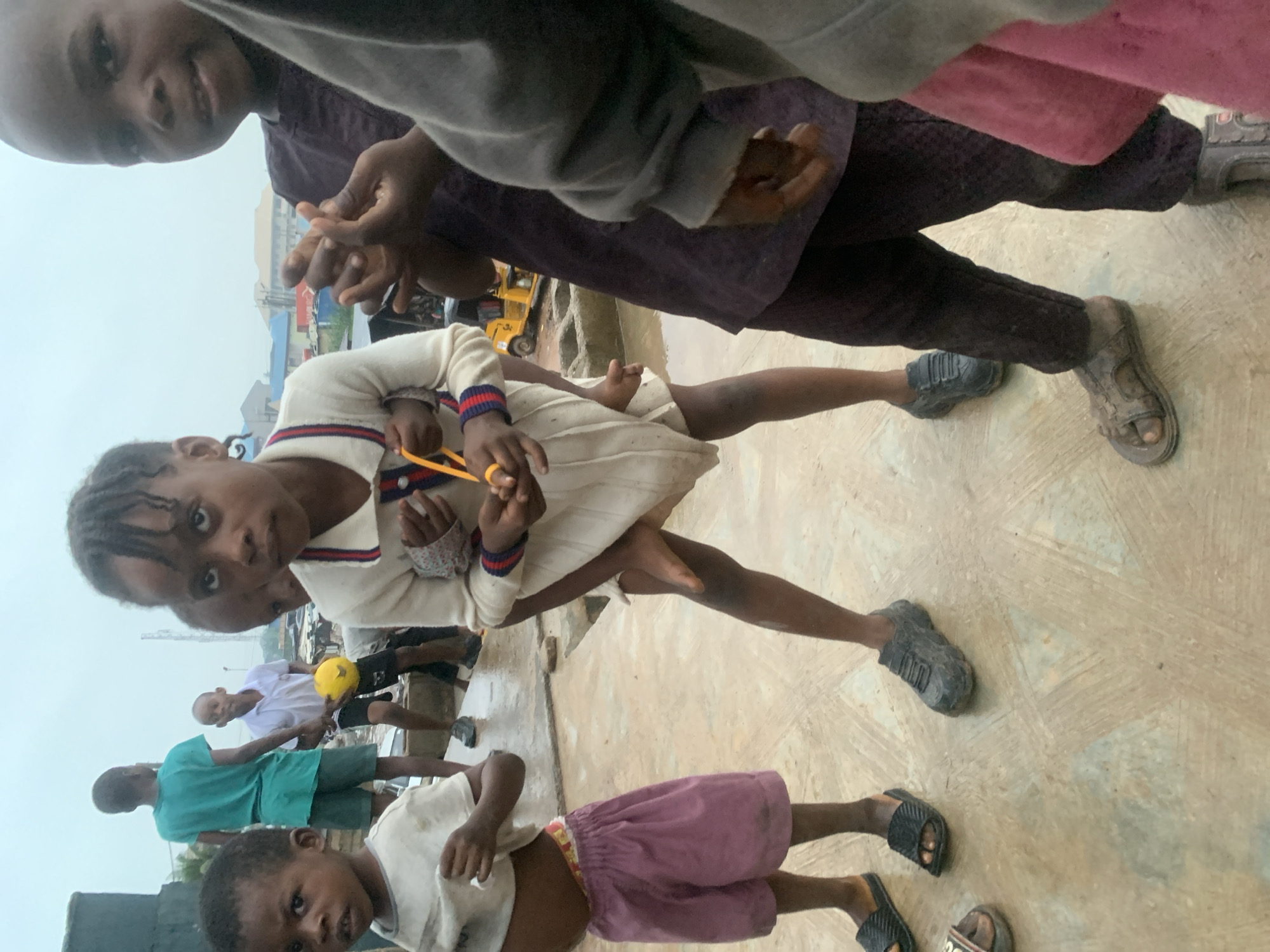 The image depicts a group of young children standing on a wet, paved surface in what appears to be an outdoor marketplace or street in Shagamu, Nigeria. The children are dressed in casual clothing, and some are holding or playing with objects. One child in the foreground is wearing a white sweater with striped trim and is holding a yellow string. Another child to the left is wearing a white t-shirt and pink shorts. In the background, other children and some structures are visible, including a yellow vehicle. The lighting suggests an overcast or rainy day. The overall impression is a candid snapshot of children in an urban environment, possibly engaged in play or daily activities.
