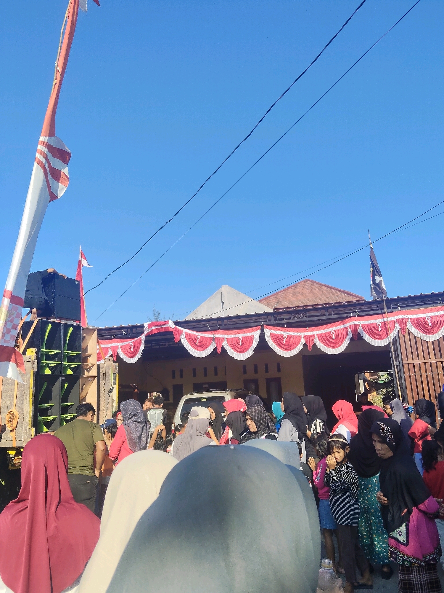 The image captures an outdoor gathering on a bright, clear day, likely in Windurejo, Indonesia, given the context. A large flagpole bearing the Indonesian flag, red and white, stands prominently on the left, partially obscuring a stack of large black speakers. A person dressed in black is seen on top of the speakers, appearing to be adjusting equipment.
In the background, a building is decorated with red and white bunting, creating a festive atmosphere. Several people are visible, many wearing headscarves, suggesting a community event. Some are standing and observing, while others appear to be interacting. A silver car is parked in front of the building.
The foreground is dominated by the back of several individuals, their heads and shoulders visible, some wearing colorful headscarves. The overall scene suggests a public celebration or a community event taking place in a residential area. The clear blue sky and the sunlight indicate it is daytime.