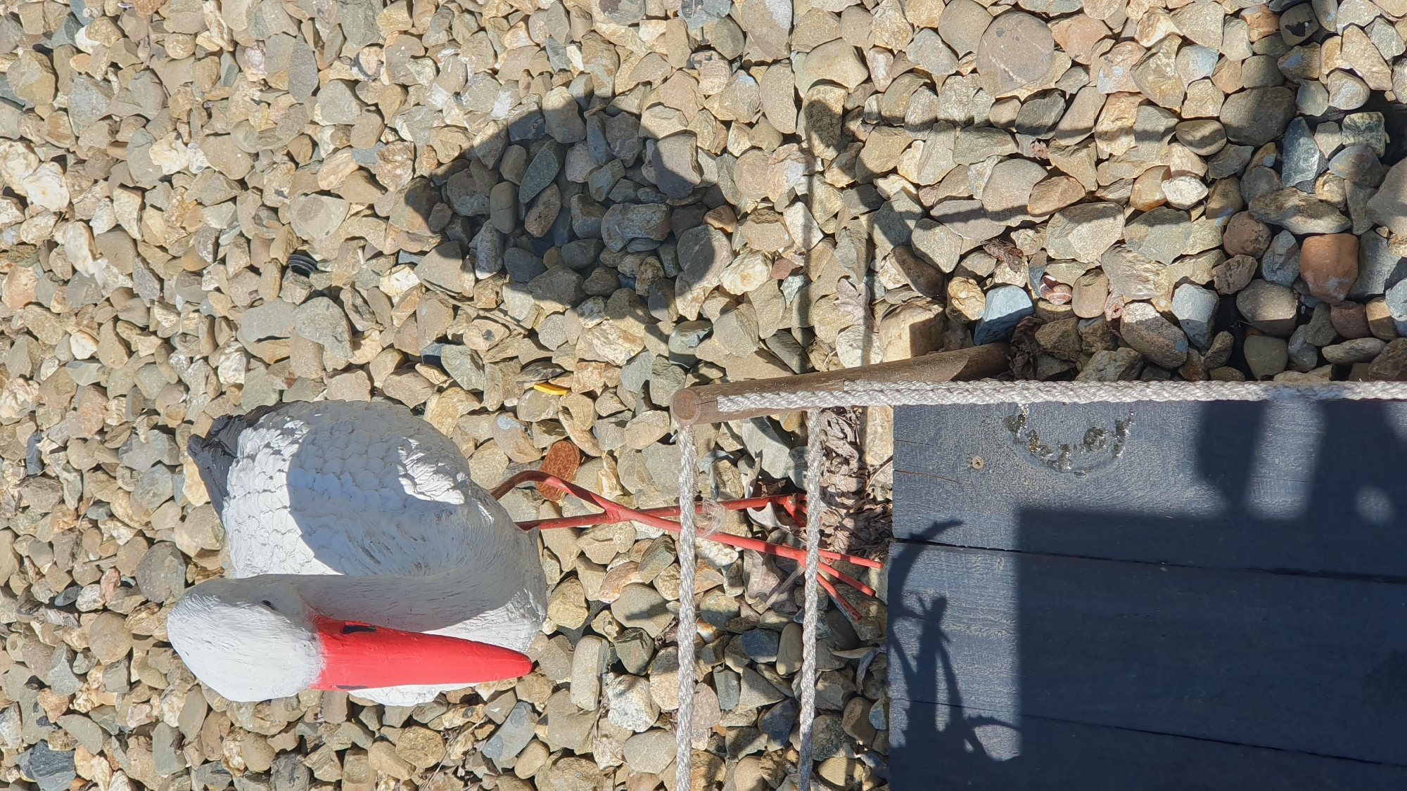 The image shows a concrete statue of a stork with a red beak and red legs, standing on a gravel surface. The stork statue is white with black markings on its wings. The background consists of various sized stones and pebbles. In the foreground, there is a dark wooden plank and a rope barrier. The scene appears to be outdoors. There are no people or actions visible in the image. The location is identified as being in the city of Ust-Labinsk, Russia.