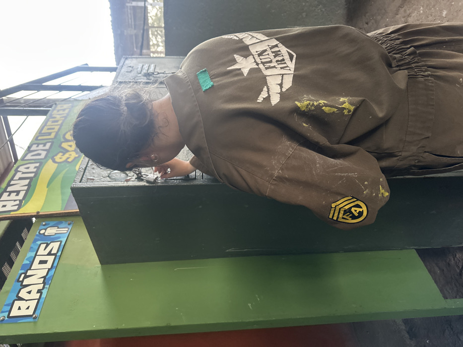 A woman wearing a tan jumpsuit is bent over, looking at a dark green storage box. The jumpsuit has patches, including a "FUN ARMY" logo on the upper back, a green square patch on the shoulder, and a yellow rank insignia on the sleeve. Signs labeled "RENTA DE LOCKER" and "BAÑOS" are visible in the background.