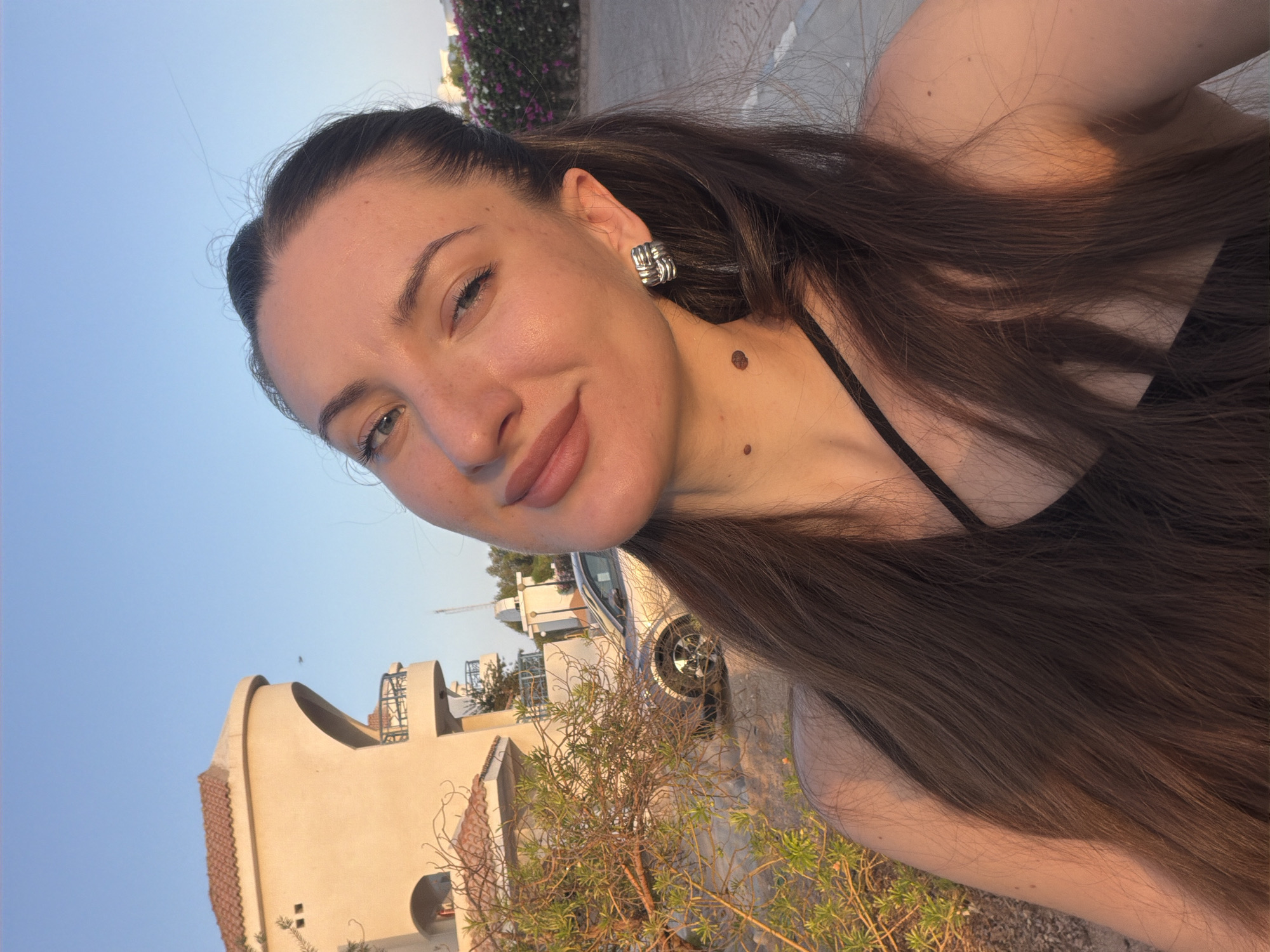 In Sharm El Sheikh, Egypt, a young woman with long, dark brown hair is captured in a close-up selfie outdoors. The setting appears to be a residential area, with a light-colored building with terracotta roof tiles and balconies visible in the background. A silver car is parked on the street, and a dry, shrub-like plant with sparse green leaves is in the foreground. The woman is wearing a black top and silver earrings. The sky is clear and blue, indicating a daytime scene, possibly during the late afternoon or early evening due to the warm, golden light illuminating her face and hair. She has a mole on her neck and another on her chest. The overall impression is a casual and relaxed moment.