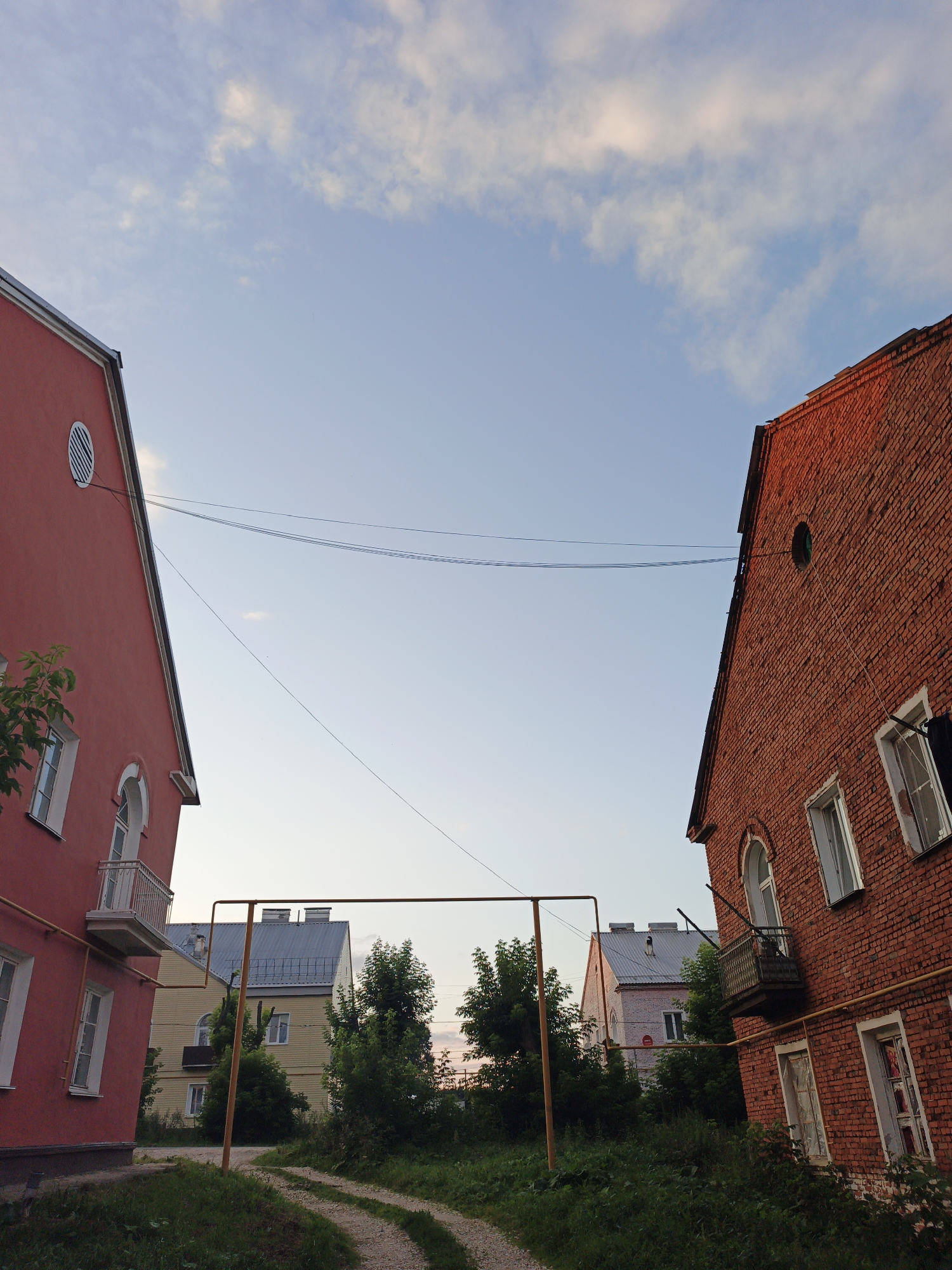 The image depicts an outdoor scene in Medvedyovka, Russia. In the foreground, two buildings frame a pathway. The building on the left is a red-painted structure with a balcony and several windows. The building on the right is constructed of red brick, also featuring balconies and windows. A yellow pipe runs horizontally between the two buildings, supported by vertical posts. In the background, more buildings are visible, including a light-colored structure with a silver roof and trees. The sky above is a pale blue with scattered clouds. The ground is covered in grass and a dirt path. The lighting suggests it is either early morning or late afternoon. There are no people or animals visible in the image.