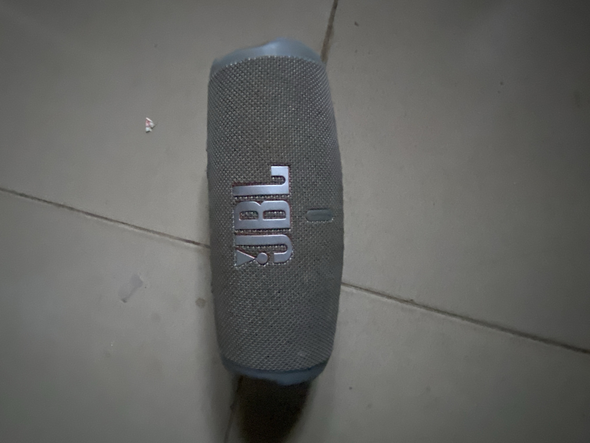 A grey JBL speaker is positioned vertically on a light grey tiled floor. Small pieces of debris are scattered around the speaker.