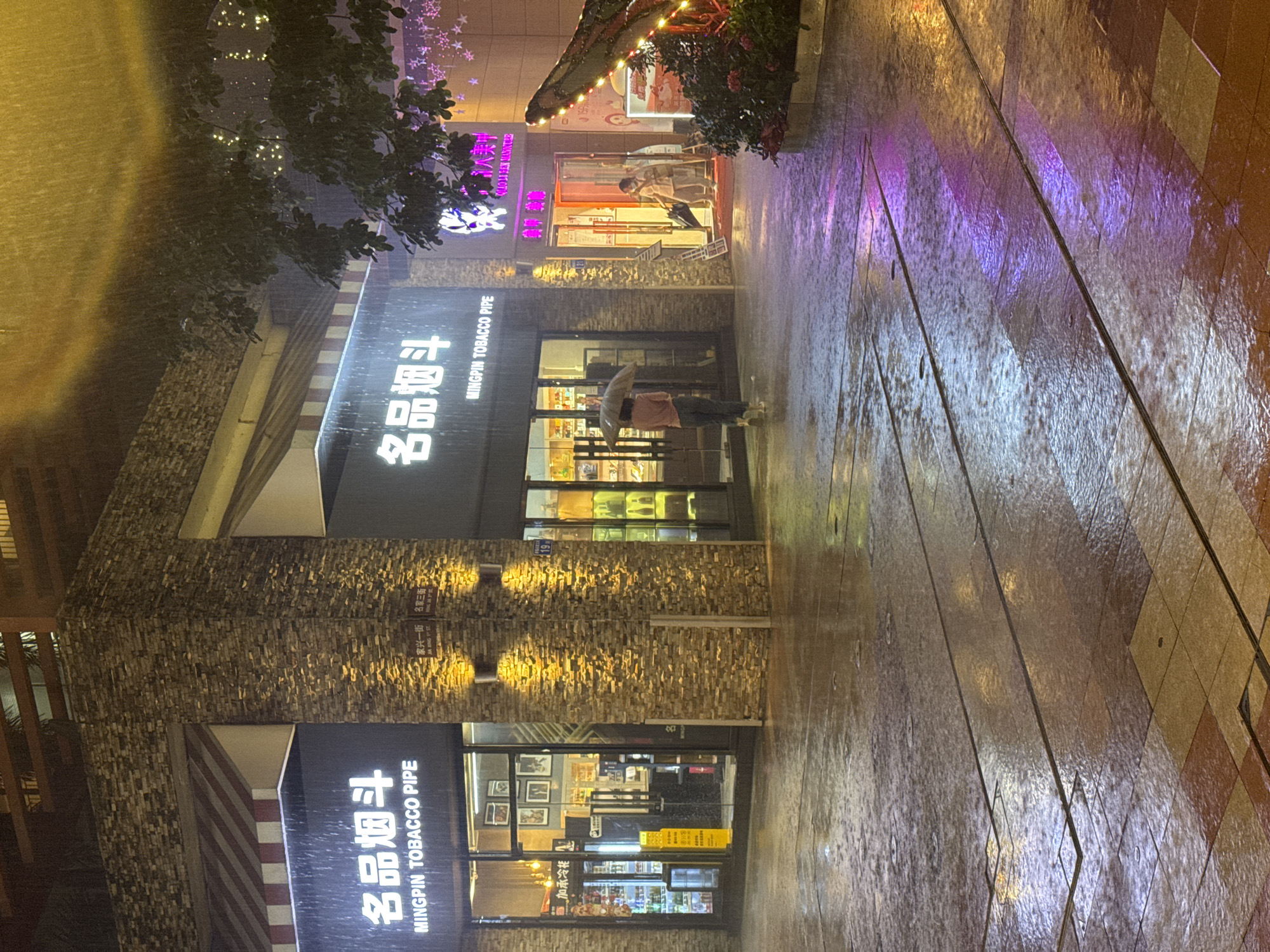 The image shows a street scene in Panyu District, China, during a heavy downpour. The view is of the storefronts of two adjacent businesses: "Mingpin Tobacco Pipe" and another business with Chinese characters. The storefronts are brightly lit, reflecting off the wet pavement. A person is visible holding an umbrella, standing in front of "Mingpin Tobacco Pipe." The sidewalk is wet, and rain streaks are visible throughout the image. Additional storefronts are visible down the street.