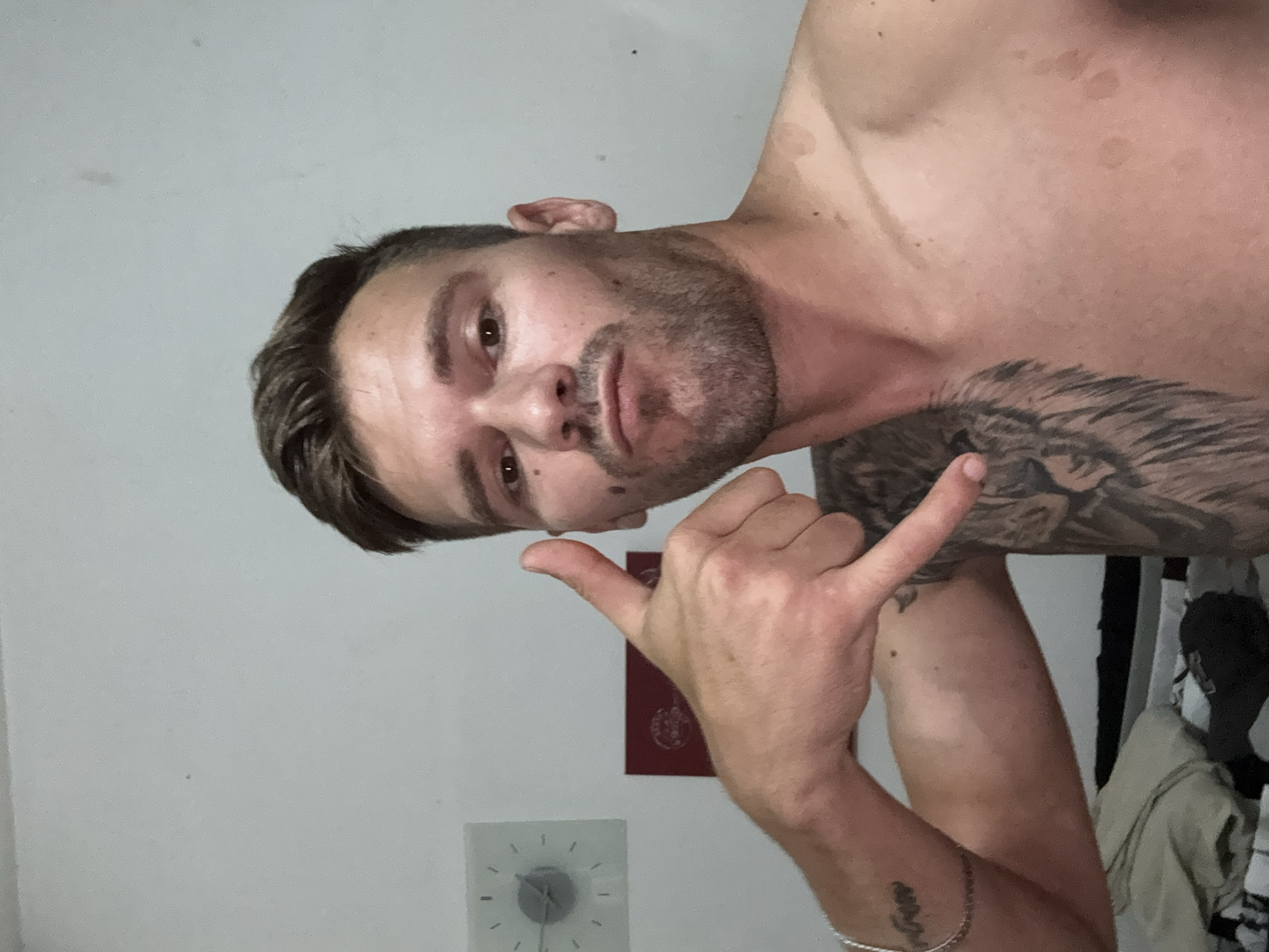 The image shows a man making a "call me" gesture with his right hand. He is shirtless and has a tattoo of a lion on his left shoulder and chest. He is wearing a bracelet on his left wrist. The background is a plain white wall with a clock on it. There are some reddish circular marks on his chest and shoulder. The man has short brown hair and a beard. He is looking at the camera with a neutral expression. The location is described as being in the city of Sesto Calende, Italy.