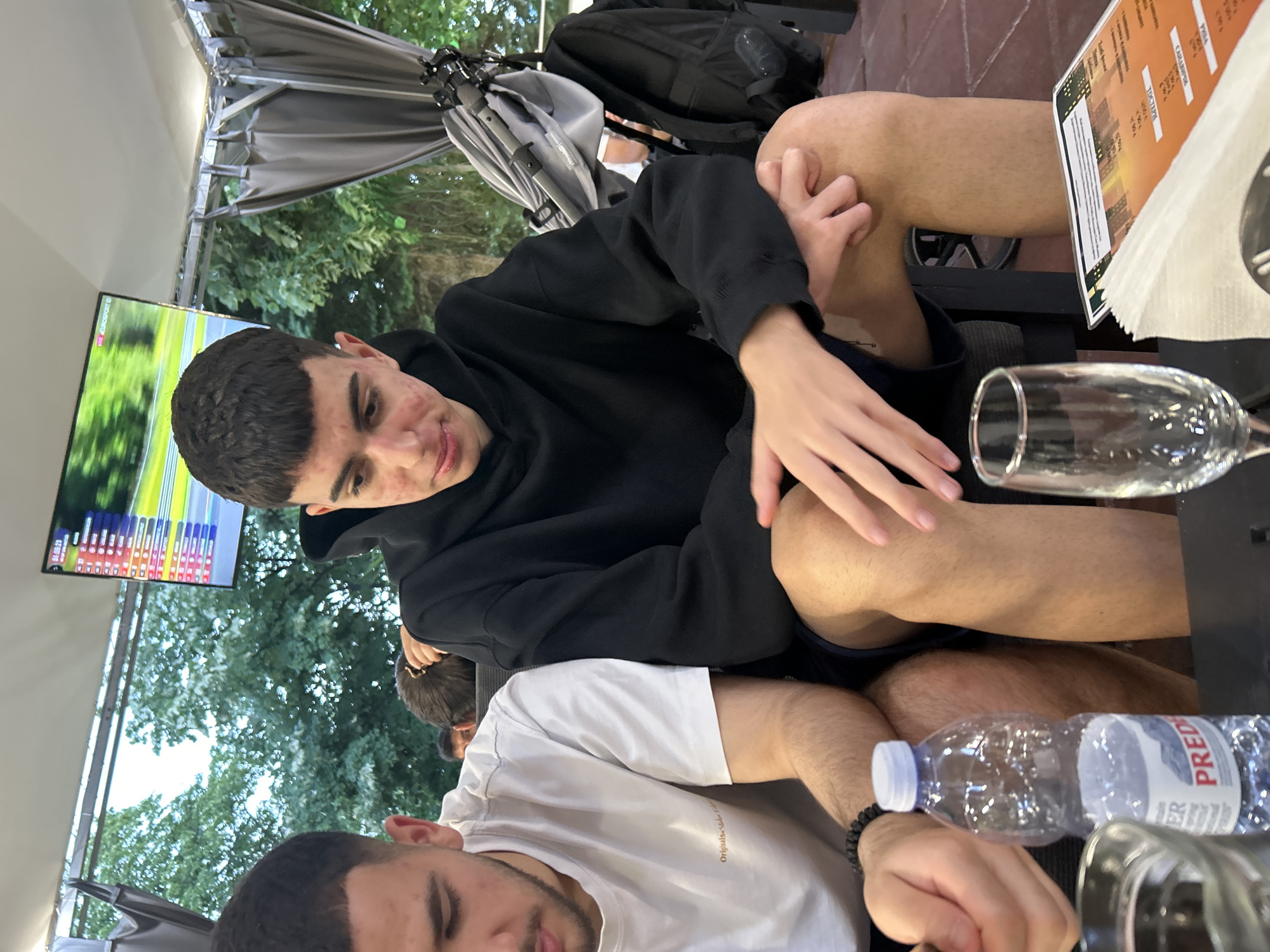 The image captures two young adult males seated at a table in what appears to be an outdoor or semi-outdoor cafe or restaurant, likely in the evening or a shaded area. The environment features a white ceiling structure with metal beams, dark grey retractable curtains or drapes on the right, and out-of-focus green foliage and trees in the background.
The individual on the left is partially visible, wearing a white short-sleeved t-shirt with "Originals..." text and a black beaded bracelet on his left wrist. He is looking downwards. On the table, a clear plastic bottle of "PREDATOR" brand water and a clear drinking glass are positioned near him.
The individual on the right is centrally framed, wearing a black hooded sweatshirt and dark shorts. He has noticeable redness on his face, consistent with acne, and is looking downwards with his hands clasped on his right knee. Adjacent to him on the table are an empty stemmed glass, a stack of white napkins, and a laminated menu featuring an orange background and text in Cyrillic script, including items such as "ТОСТЕР" (TOASTER), "САЛАТИ" (SALADS), and "ПИЛЕ" (CHICKEN).
In the background, a flat-screen television displays a sports broadcast, identified by a "EUROSPORT" logo, a timer reading "01:08:23", and a list of names and data, indicative of a racing event scoreboard. To the right of the television, a folded grey baby stroller and a black backpack are present. The floor is tiled with reddish-brown terracotta-like tiles. While the menu contains Cyrillic script, no specific visual information within the image definitively identifies the location as Blagoevgrad, Bulgaria.