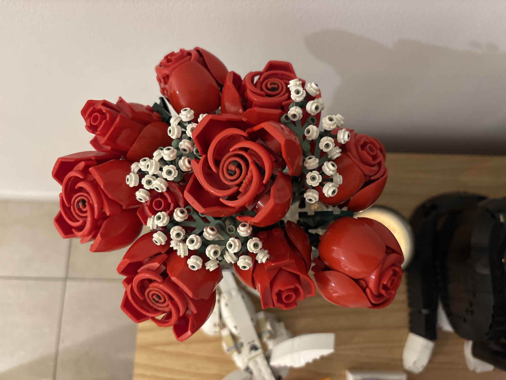 The image shows a bouquet of red Lego roses with white Lego baby's breath flowers, viewed from directly above. The bouquet is on a light-colored wooden surface. Partially visible beneath the bouquet is a white Lego model of a spaceship or vehicle. To the right of the bouquet, a black and white Lego figure, possibly a penguin or other animal, is visible. The environment appears to be indoors, likely a home. There is no indication of a specific location within Buenos Aires, Argentina, or any other location.