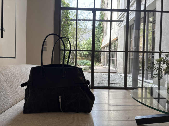 A dark-colored tote bag with black leather top handles and a textured lower body featuring a vertical front zipper and side straps rests on a light beige, woven sofa. To the right, a circular glass coffee table with a dark base displays a vase containing green foliage. The scene is viewed through a large, multi-paned industrial-style window with dark frames, which overlooks a private inner courtyard. The courtyard features a light gravel ground covering leading to a wooden deck or patio. Beyond, a building facade of light-colored concrete or stone is visible, partially covered in ivy, with additional windows. The architectural style and private courtyard environment indicate a location within the city of Paris, France. No people or active interactions are present.