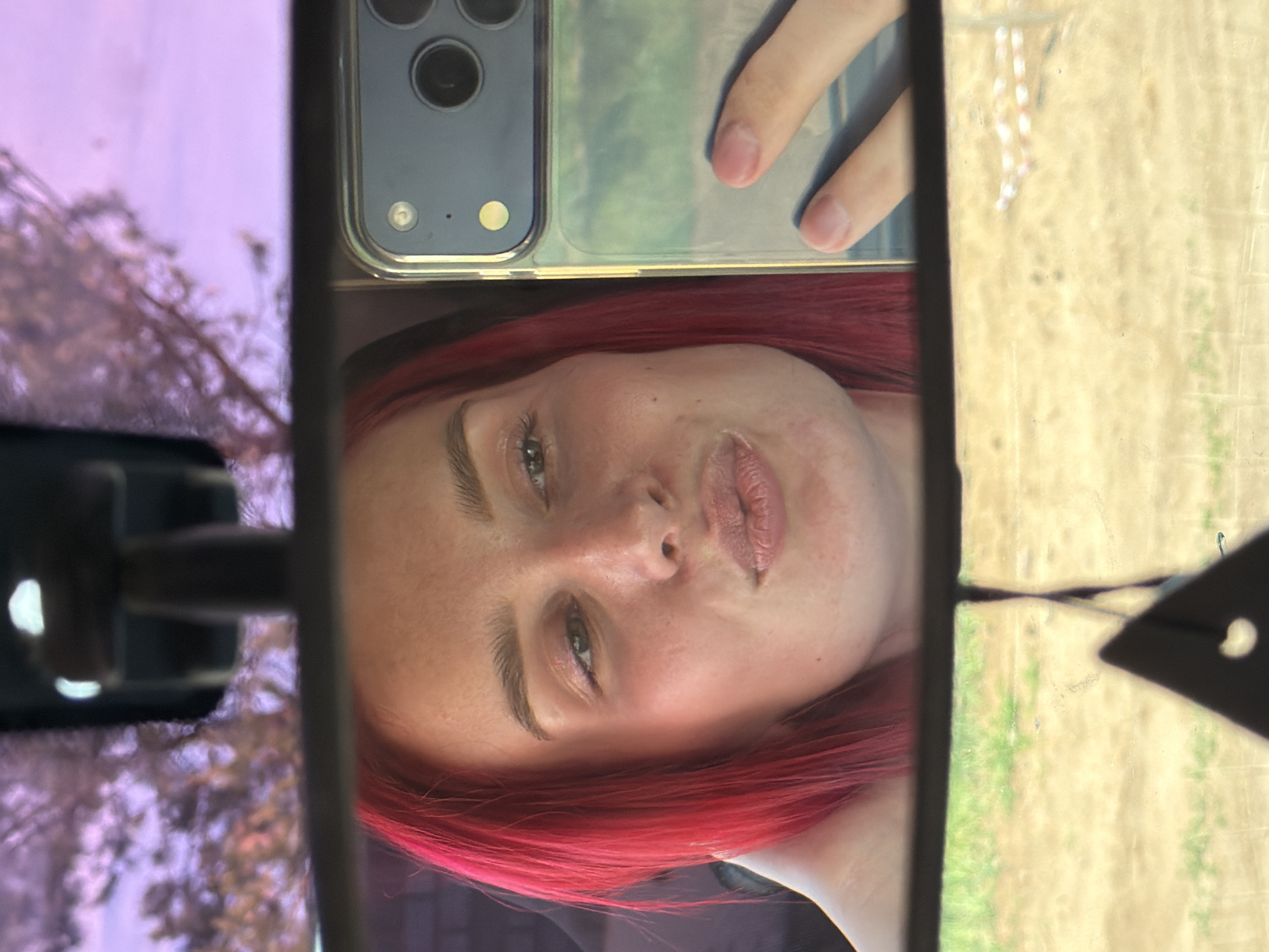 A woman with red hair is taking a selfie in a car's rearview mirror. She is holding her cell phone, a blue iPhone with multiple lenses, in front of the mirror. The scene visible in the rearview mirror includes a blurred exterior view. The specific location in the city of Chaikovsky, Russia, cannot be determined from the image.