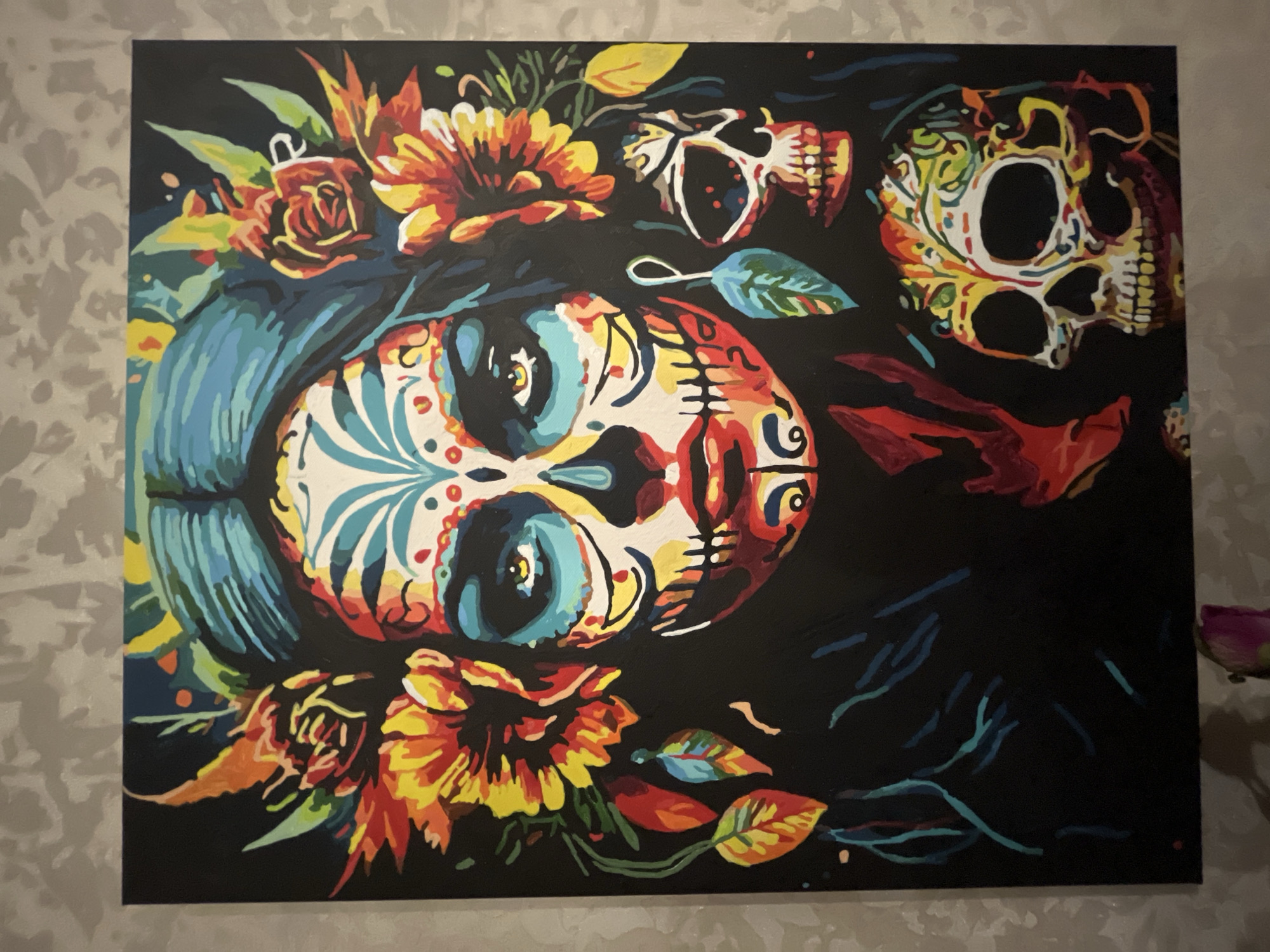 The image shows a painting of a woman adorned with Dia de los Muertos makeup, featuring a skull motif. She is surrounded by colorful flowers and leaves. Two skulls are depicted; one is positioned to the right of her face, appearing as an earring or accessory, and another larger skull is shown below her face. The overall aesthetic is vibrant and decorative, typical of the Sugar Skull tradition. There is no indication of its location within the city of Bryansk, Russia.