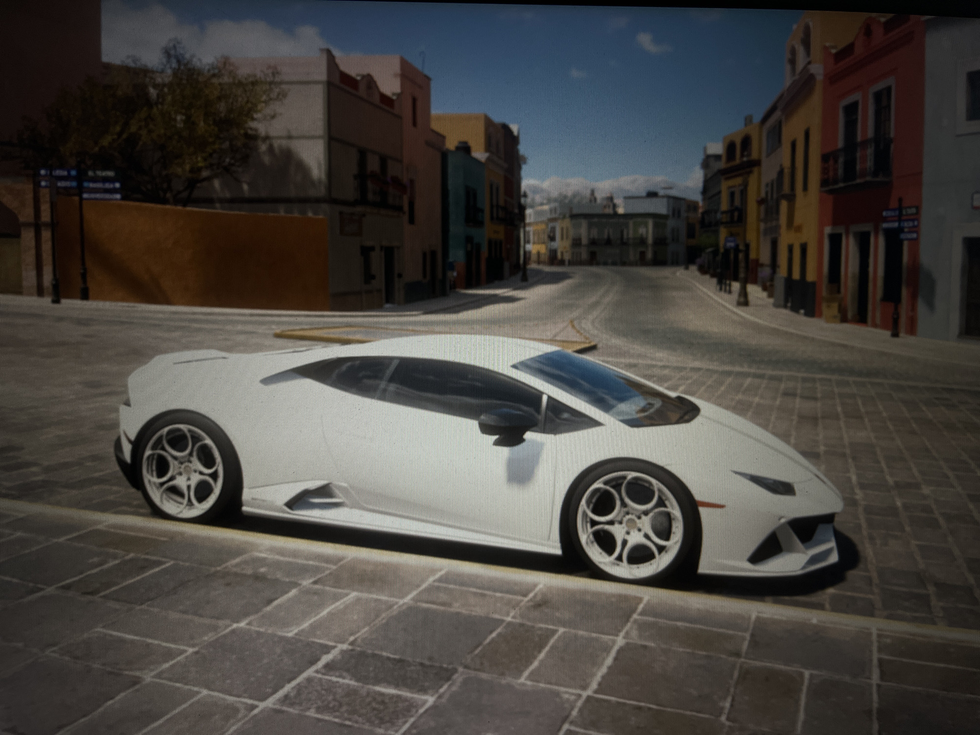 The image depicts a white Lamborghini Huracán parked on a cobblestone street in a city. The street is lined with colorful buildings, featuring balconies and architectural details consistent with a European or Latin American urban setting. The sky is blue with some scattered clouds. The car is the primary subject, with its sleek design and distinctive wheels prominently displayed. The environment appears to be a well-maintained city square or street, suggesting a scenic and potentially tourist-frequented location. There are no people visible in the image. The image quality is slightly pixelated, suggesting it may be a photograph taken from a screen or a low-resolution capture. The location is not Varna, Bulgaria.