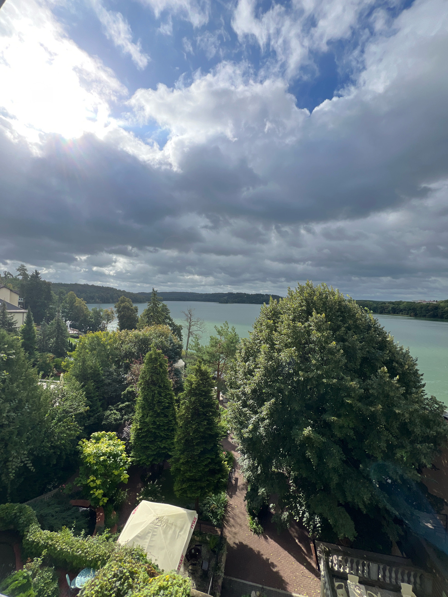 The image depicts a scenic view of a lake and its surrounding greenery, likely from an elevated vantage point. A large, calm body of water stretches into the distance, bordered by lush green trees and foliage. The sky above is a dramatic mix of bright, sunlit clouds and darker, more ominous clouds, suggesting a dynamic weather system.
In the foreground, various trees and bushes create a dense, verdant landscape. Some buildings are partially visible among the trees on the left side of the image. A pale yellow or white umbrella is set up in a garden area, along with some outdoor furniture, suggesting a recreational or hospitality setting. The ground in the foreground appears to be paved with brick.
The overall impression is of a natural landscape with some human presence, possibly a park, a resort, or residential area near a lake.
This view is consistent with many locations in Mrągowo, Poland, a town known for its numerous lakes and forested surroundings, offering similar vistas. For instance, the view could be from a hotel or a private residence overlooking Lake Czos or another of the town's many picturesque water bodies.