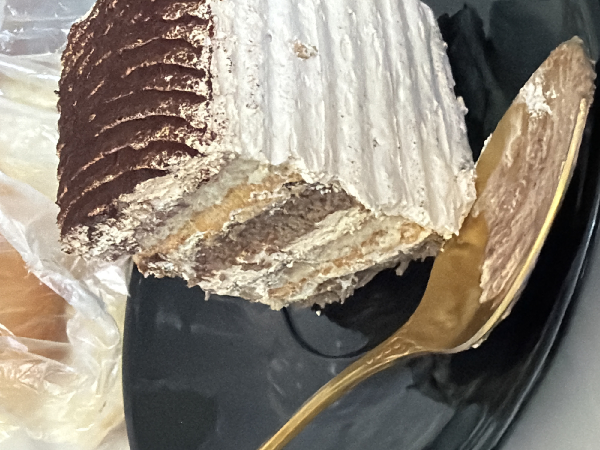 A slice of tiramisu cake is presented on a black plate, with a golden spoon resting on it. The cake has layers of cream and sponge, dusted with cocoa powder on top. The background shows a crumpled plastic bag. The image is a close-up shot, with the cake and spoon occupying most of the frame. There is no indication of location in the city of Strășeni, Moldova.