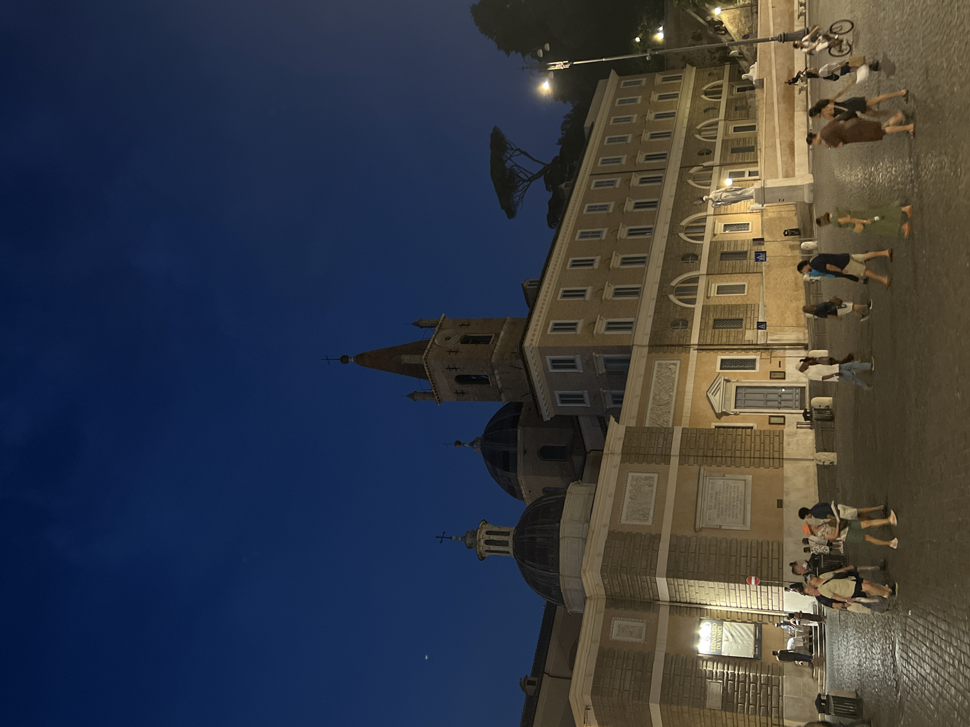 This is a nighttime, eye-level, full-shot photograph of a paved plaza in front of a large building complex in Rome, Italy. The building features a prominent clock tower with a red, conical spire and a bell tower. To the left of the clock tower are two domes, one smaller and lighter in color, the other larger and darker. The main building appears to be an ecclesiastical or historical site, with classical architectural elements such as arched windows and sculpted friezes. The sky is a deep, clear blue, suggesting twilight or early evening.
Several people are walking across the plaza. They are dressed in casual summer clothing. Some are walking individually, while others are in small groups, appearing to be tourists or locals. The cobblestone pavement of the plaza is illuminated by artificial lighting from street lamps and the building itself. To the right of the building, a tall, distinctive pine tree stands on a small hill. In the distance, on the right side of the frame, a street lamp is visible, with light emanating from it. The scene conveys a sense of calm evening activity in a historic urban setting.