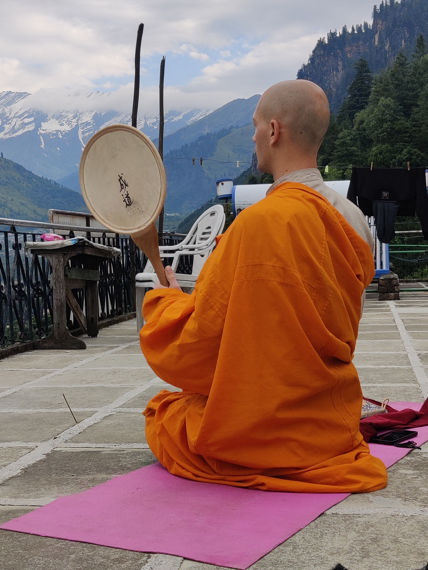 A bald, fair-skinned monk wearing an orange robe sits cross-legged on a pink yoga mat on a tiled rooftop in Vashisht, India. He holds a circular drum with Chinese characters on it, supported by a wooden handle. The drum faces away from the camera, revealing the characters on its surface. In the background, snow-capped mountains and lush green trees are visible under a cloudy sky. A black jacket and other garments are hung on a clothesline to the right of the frame. A wooden table with a black metal railing is positioned to the left of the monk.