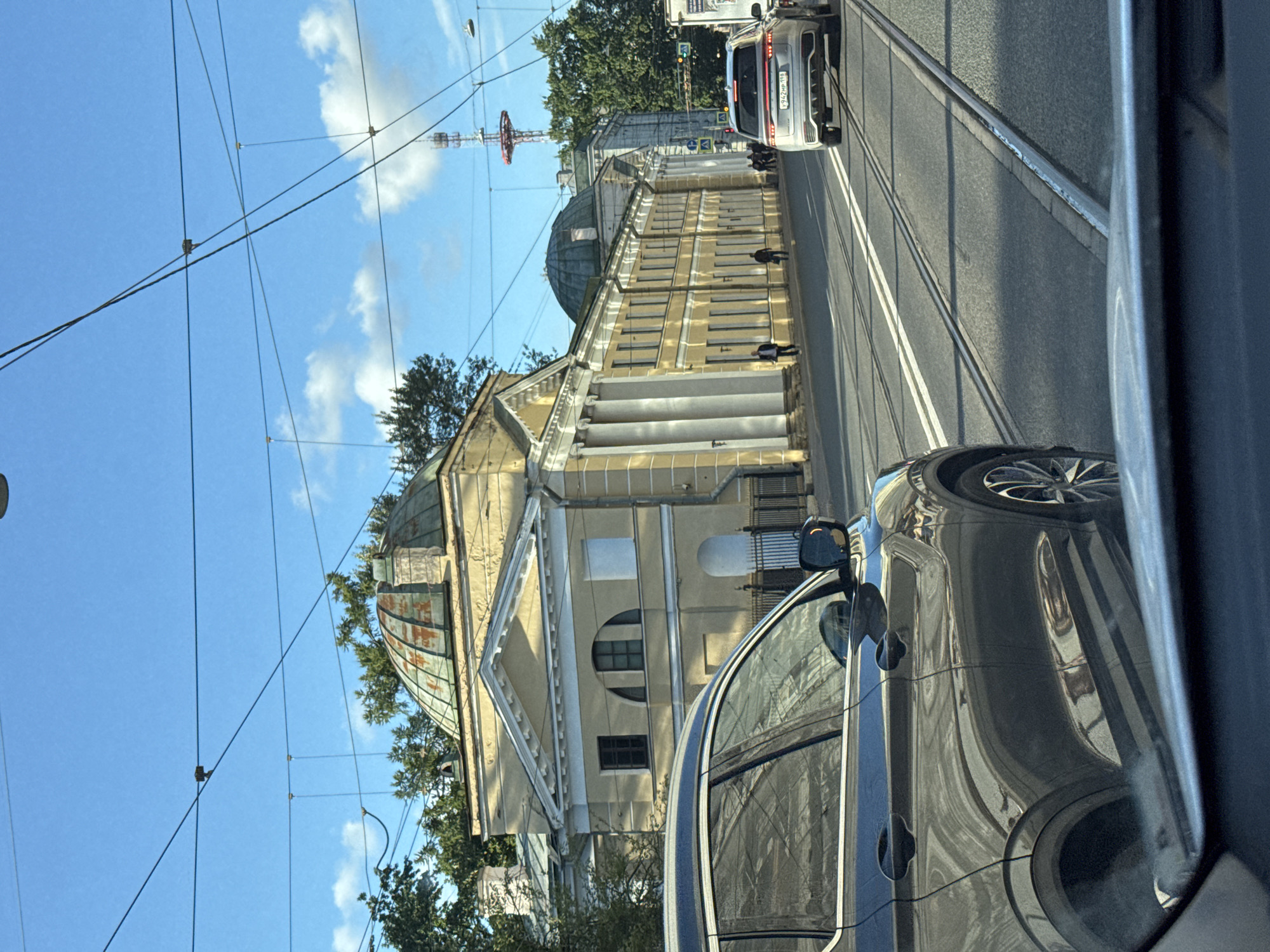 The image depicts a street scene in Saint Petersburg, Russia, under a clear blue sky with scattered clouds. In the foreground, the side of a dark gray SUV is visible, reflecting the surrounding environment. The SUV is on the right side of the road, next to tram tracks. Further down the street, another silver SUV is seen, with its rear license plate readable as "У942МР 198".
On the left side of the street, a long, pale yellow neoclassical building with white columns lines the roadway. The building features a prominent dome on its left side and a pedimented facade with arched windows. Trees and foliage are visible behind and around the building. A tall radio tower with red structural elements stands in the background on the right side of the image. Several individuals are walking on the sidewalk in front of the yellow building. The presence of tram tracks indicates it is a city with a functioning tram system. The overall atmosphere suggests a daytime setting in an urban environment.
