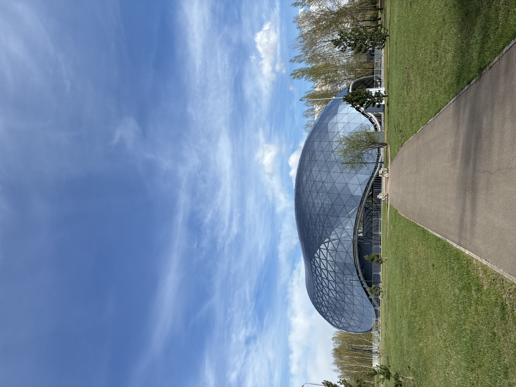 A vertical, eye-level outdoor shot captures a building with a unique, curvilinear glass facade. The building is situated along a pedestrian path that stretches from the lower right corner towards the center. Adjacent to the path is a green grassy area, interspersed with trees. The sky, taking up the majority of the image, is blue with streaks of white clouds. The building's architecture is a standout feature, resembling a dome or a shell, constructed from triangular glass panels. The location appears to be a park or recreational area within an unidentified city.