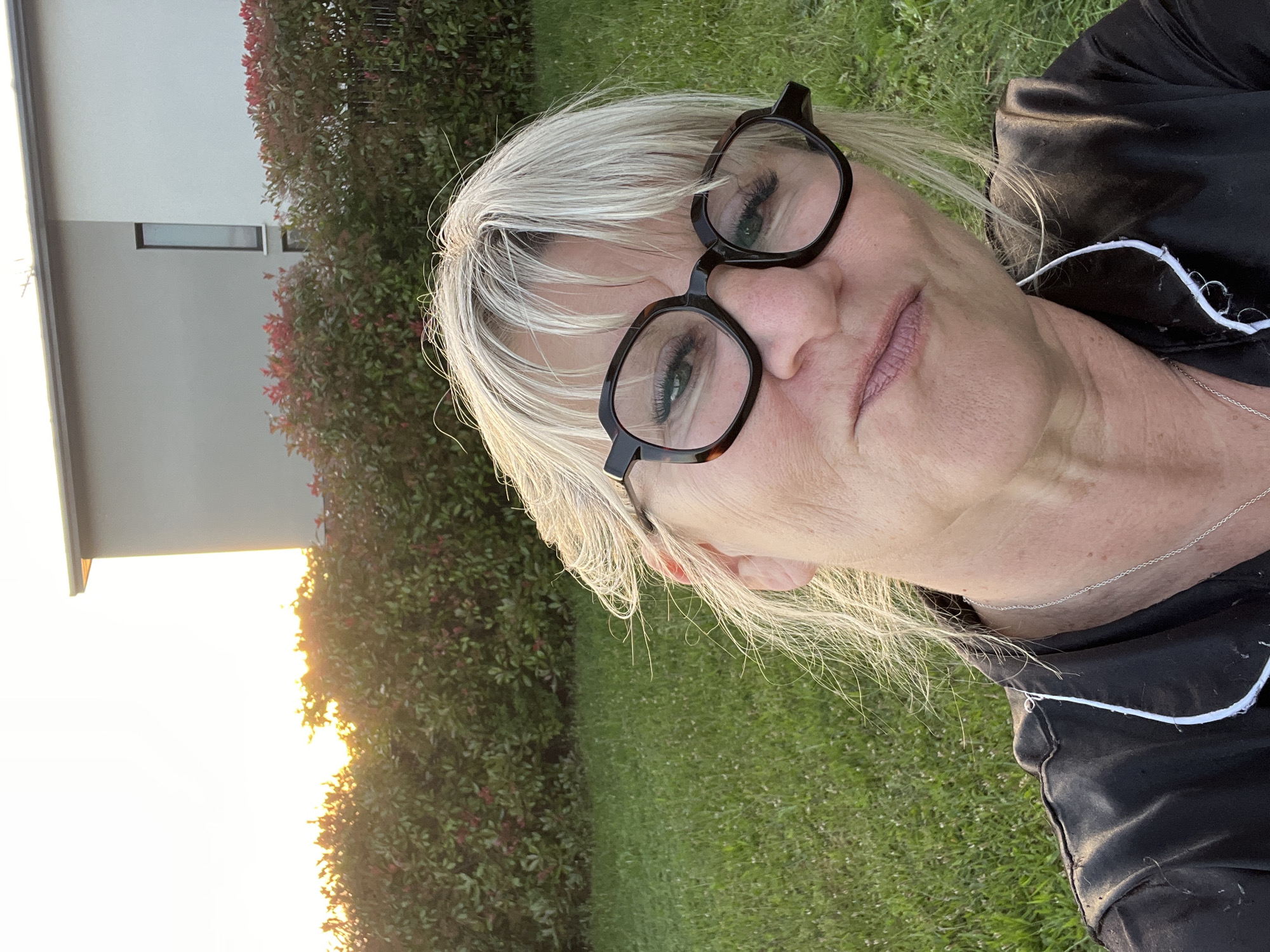 The image is a close-up selfie of a mature woman with fair skin and blonde hair, wearing black-framed glasses and a dark-colored shirt with white piping. She is looking directly at the camera with a slight smile.
The background includes a building with a light-colored facade and a small window. Adjacent to the building is a dense hedge with green and reddish foliage. The lower portion of the frame features a grassy area, and there is a bright glow of sunlight visible in the distance, likely indicating either sunrise or sunset. Based on the provided information, the location is Chambœuf, France.