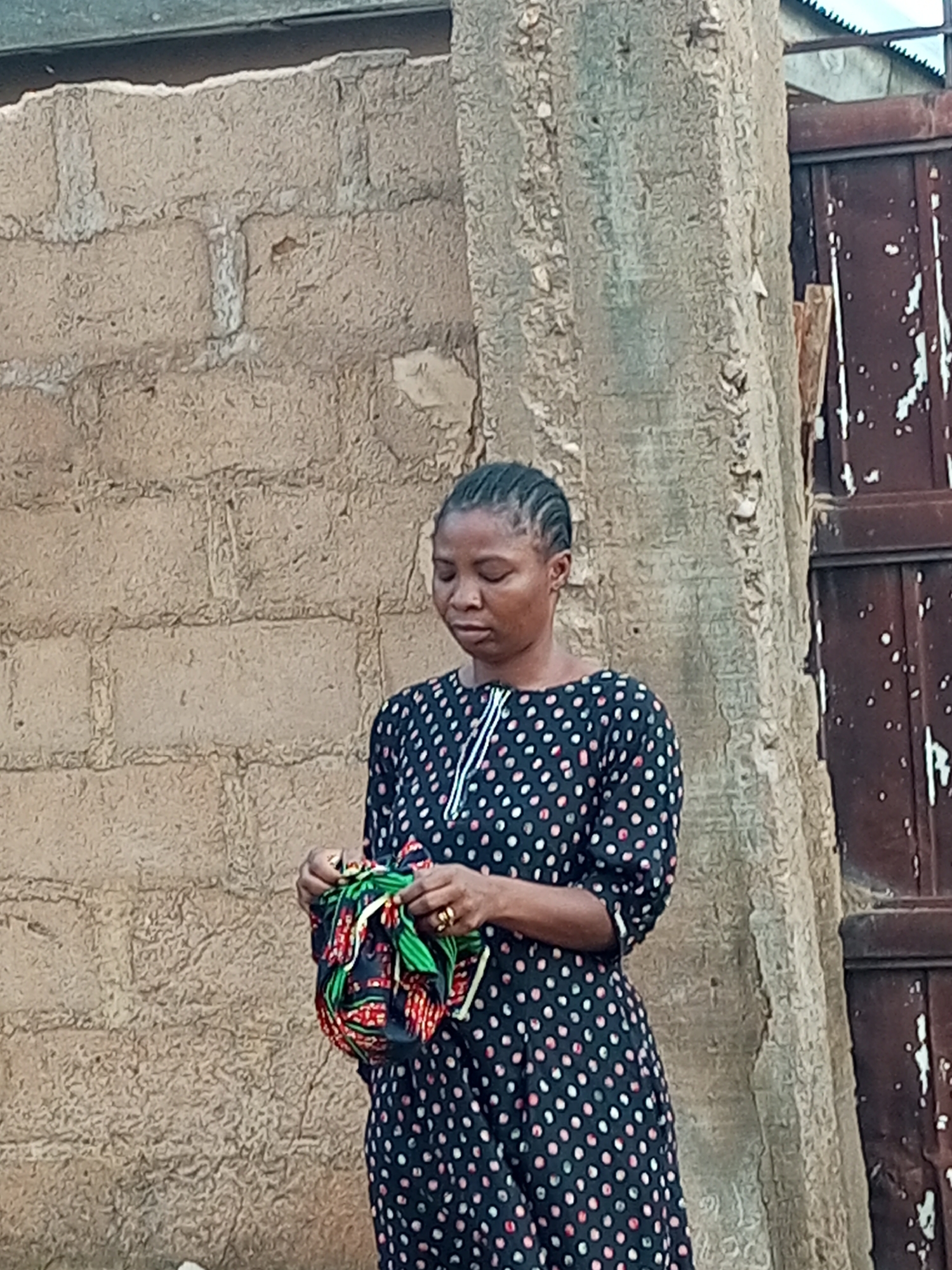 An exterior, eye-level, medium shot captures a woman standing in front of a rough, light brown brick wall and a rough, grey concrete pillar. The woman, with her dark hair braided back, is wearing a dark, long-sleeved dress adorned with white polka dots and what appears to be a thin white stripe around the neckline. Her hands are occupied with a small, colorful fabric bundle. The woman is facing slightly to her right, with her gaze directed downwards, seemingly focused on the object in her hands. To the right of the pillar, a weathered, dark brown metal door is visible, with peeling paint creating a streaked effect. The overall impression is of a candid moment, possibly in an outdoor marketplace or residential area in Tempure, Nigeria. The lighting is somewhat diffused, suggesting it could be daytime.