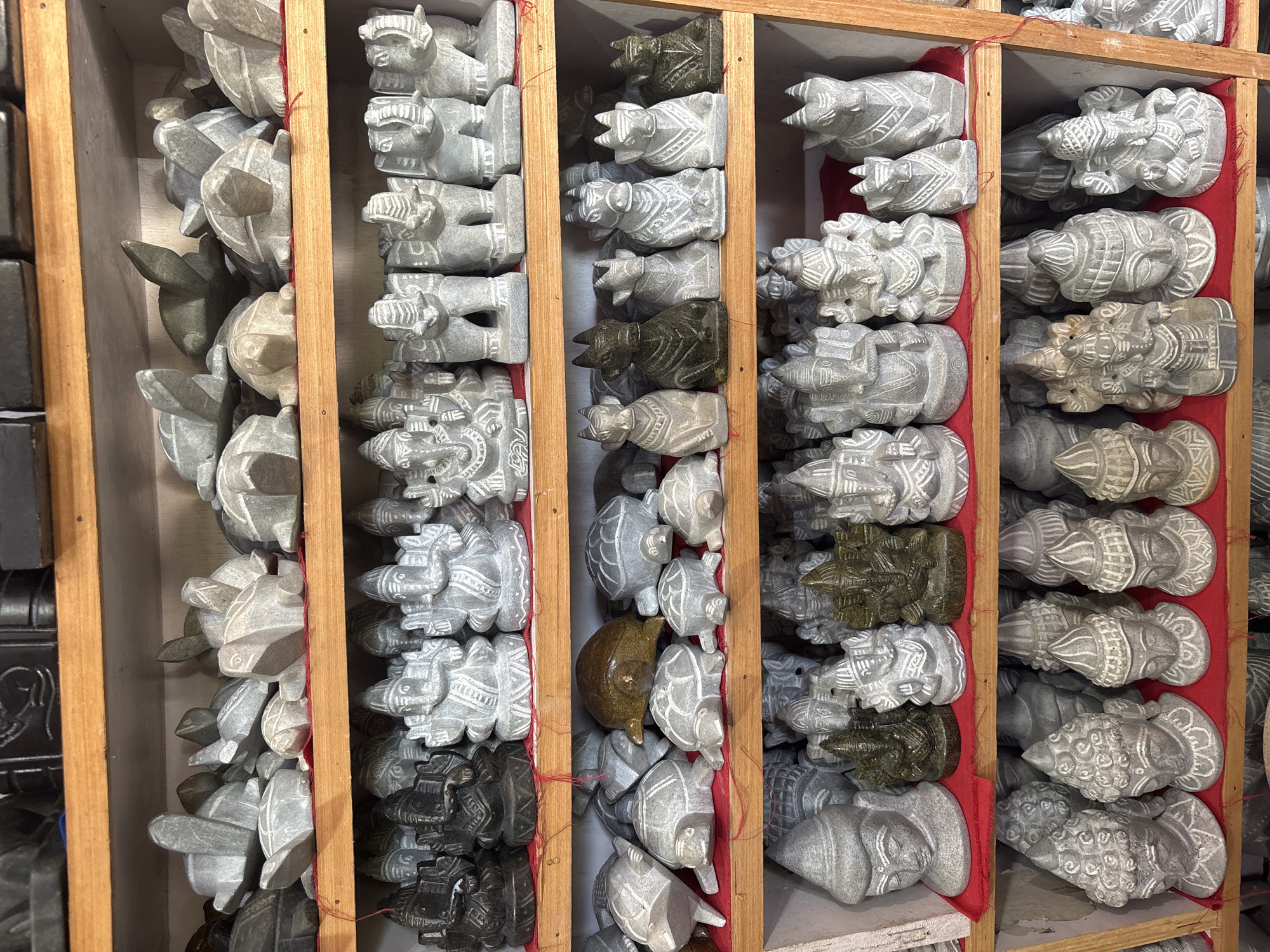 The image shows a wooden display shelf filled with small, carved stone figurines. The shelf is divided into several compartments, each containing multiple figurines. The figurines appear to be of various animal shapes, including turtles and oxen. The figurines are primarily grey. Some are light green-grey or dark. Some of the compartments have red cloth lining the back. The location is Kamalapura, India.