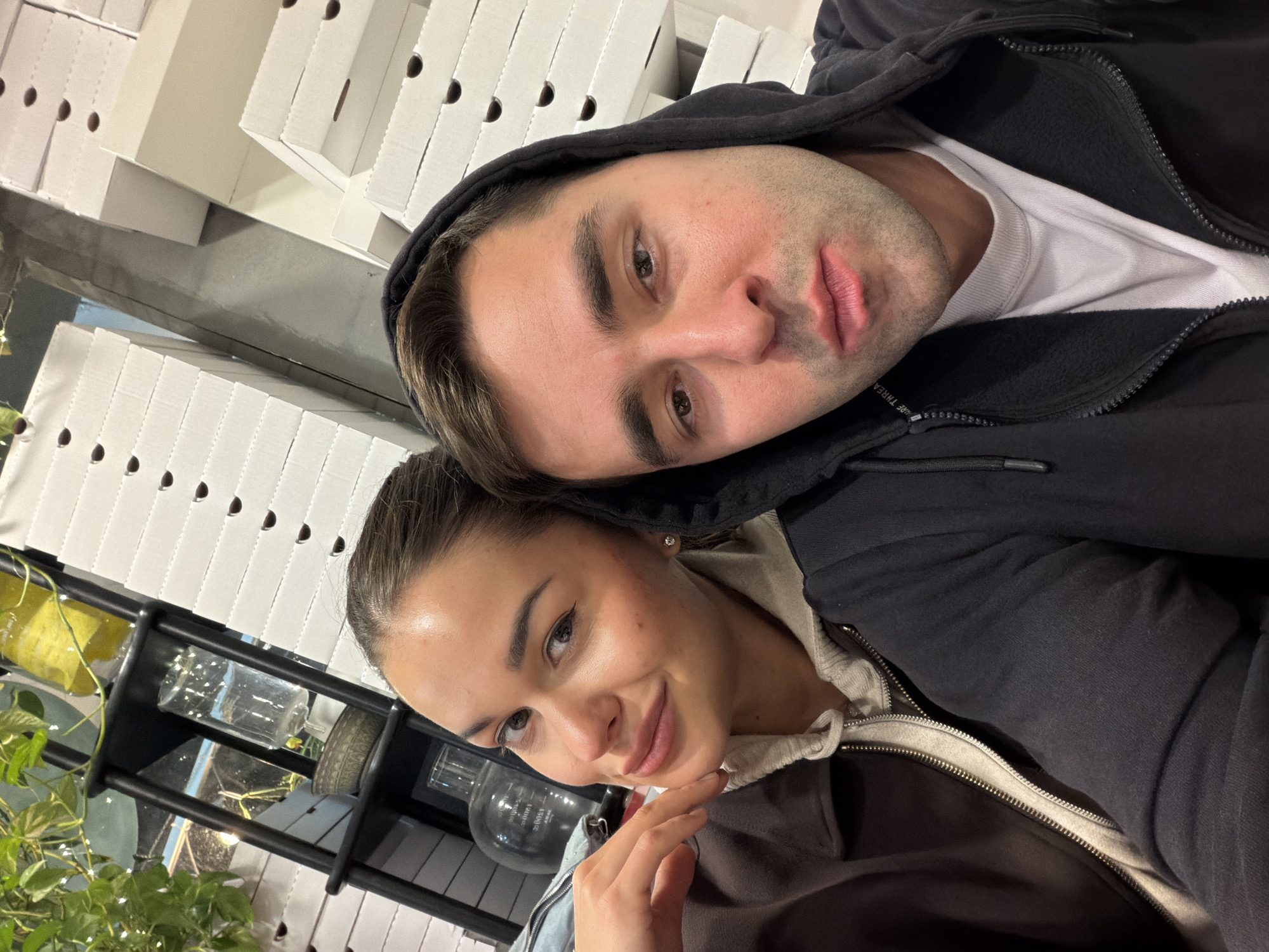 The image depicts two individuals, an adult female and an adult male, captured in a close-up, selfie-style photograph within a commercial establishment in Tel-Aviv, Israel.
The female, positioned on the left, has light skin, dark hair pulled back, and light makeup including winged eyeliner. She wears a dark brown collared jacket over a light grey zipped hoodie. Her right hand is raised, with fingers gently resting below her chin, and she displays a subtle smile while looking directly at the camera. A small stud earring is visible on her right earlobe.
The male, positioned on the right, has light skin, dark hair, and visible stubble. He is wearing a black hooded sweatshirt with the hood up, over a white crew-neck t-shirt. His expression is a slight pout, and he also looks directly at the camera.
The background primarily features numerous stacks of plain white cardboard boxes, consistent with pizza boxes or similar food packaging, suggesting the venue is a pizzeria or a restaurant offering extensive take-out services. To the far left, behind the female, a black multi-shelf unit is visible, displaying green foliage with small lights, a large clear glass jar containing yellow material, a clear glass decanter with liquid, and a decorative patterned bowl. The lighting appears to be artificial, indicative of an indoor setting.