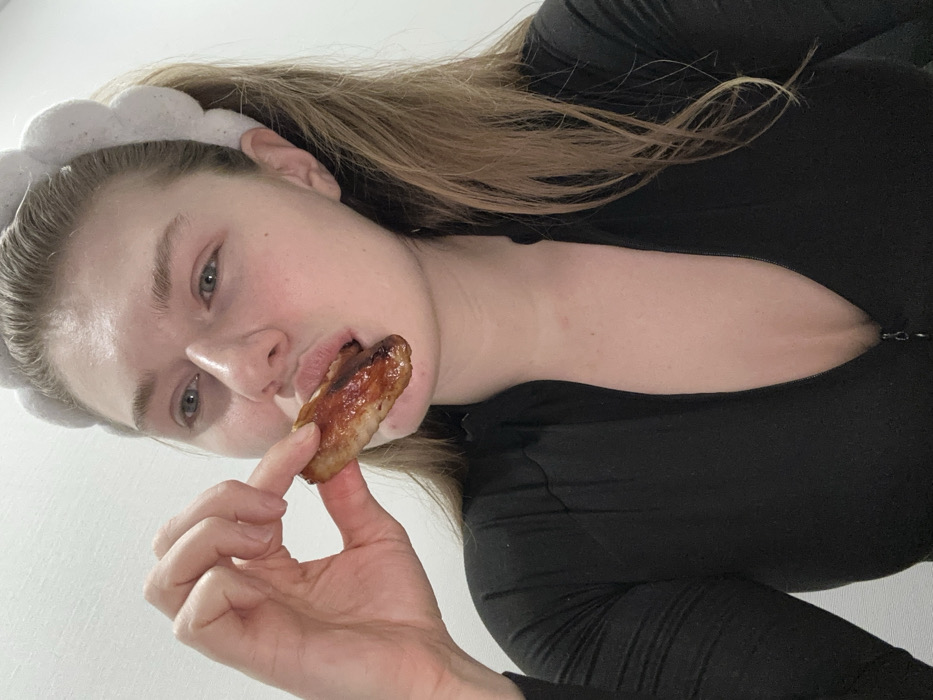 The image captures a close-up, high-angle shot of a person, likely female, engaging in an act of eating. The person is positioned diagonally across the frame, looking directly into the camera.
The subject has fair skin, light-colored eyes (appearing blue or green), and light brown or blonde hair that falls over their shoulders and back. They are wearing a fluffy, light-colored (possibly white or light grey) headband. Their expression is direct, with eyes wide open and lips slightly parted around a piece of food. There's a slight natural sheen on their skin, particularly on the forehead and cheeks.
They are wearing a black, long-sleeved garment, possibly a top or a dress, which features a zipper detail at the front that is partially undone, creating a deep V-neckline.
In their right hand, they are holding a piece of cooked meat, which appears to be a chicken wing or drumette, glazed with a dark, glistening sauce. The meat is positioned with one end partially inserted into their mouth, indicating they are either taking a bite or have just taken one. Their fingers are wrapped around the other end of the chicken piece.
The background is a plain, solid light color, likely a white or off-white wall, providing no additional context about the location. The overall lighting is bright and even. The composition is dynamic due to the diagonal alignment of the subject within the frame. No text is visible in the image.