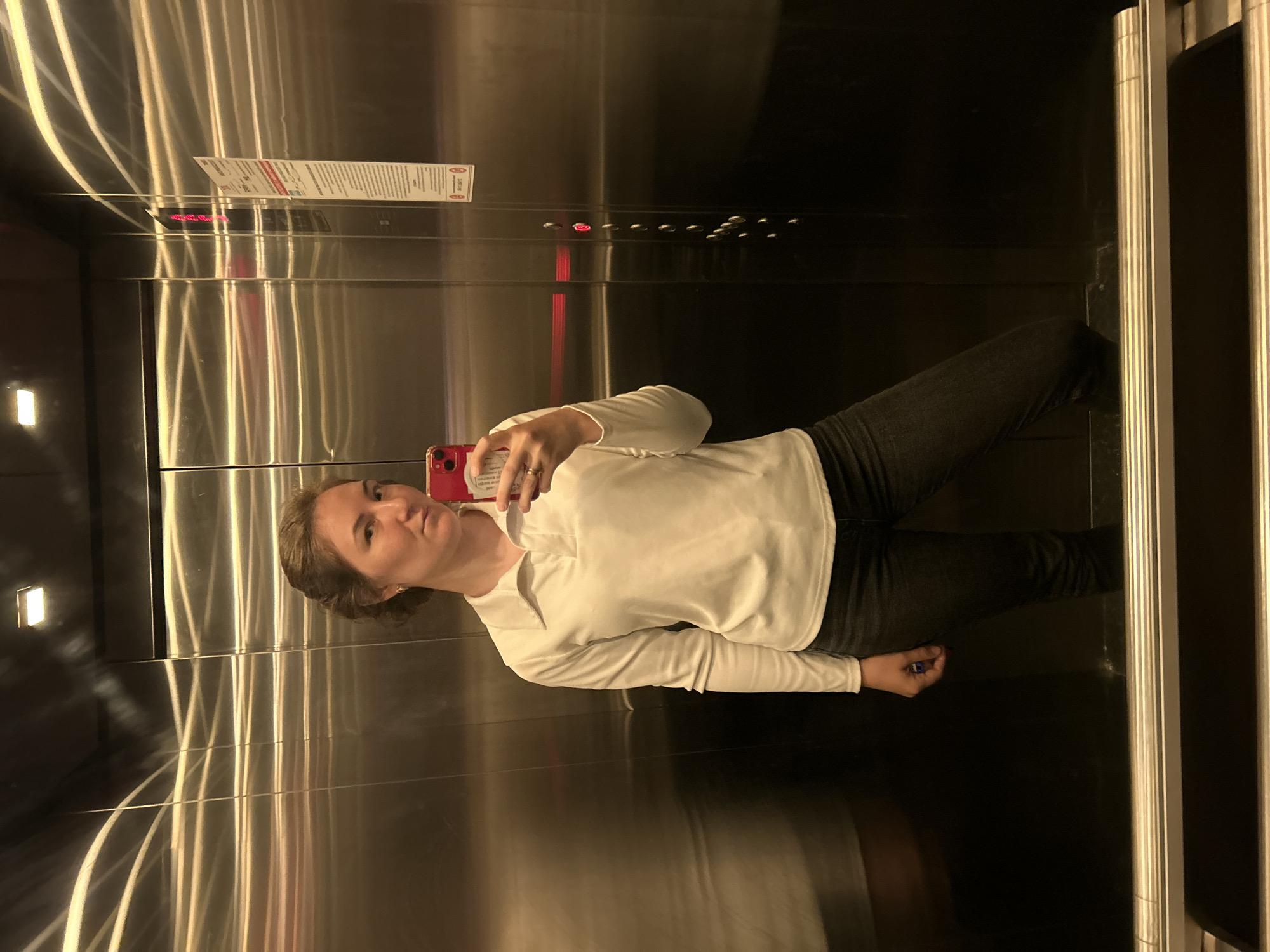 A woman is standing in an elevator, taking a selfie with a red smartphone. The elevator has brushed metal walls with linear patterns and a control panel with buttons. A rectangular informational sign is visible above the buttons. The woman is wearing a long-sleeved white shirt and dark pants. The location is in Noviy, Russia.