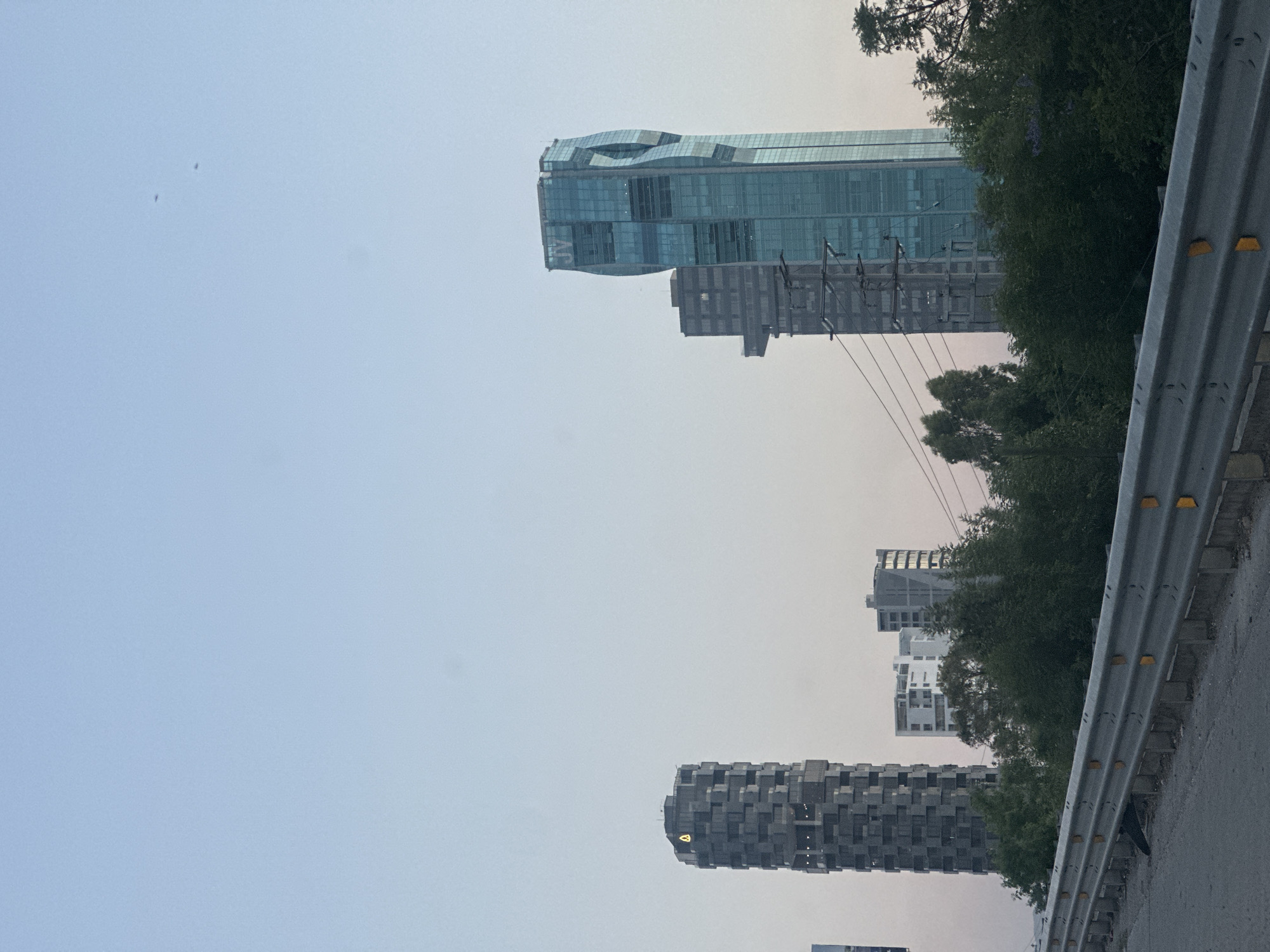 The image captures an urban landscape under a pale blue to light grey sky, suggesting either early morning, late afternoon, or an overcast day. On the right side, a tall, modern skyscraper dominates the view, featuring a distinctive wavy, blue-green glass facade. The letters "JV" are visible on one of its upper sections. Below this skyscraper, utility poles and power lines crisscross, partially obscured by trees and other buildings.
On the left side of the frame, another prominent building stands, characterized by a dark, blocky, or textured facade. Near its top, a small yellow emblem or light is visible. In the mid-ground, several other grey and white buildings of varying architectural styles are nestled among scattered trees and dense foliage.
A metal guardrail with yellow rectangular reflectors runs along the immediate right edge of the image, indicating the presence of a road or an elevated structure. Two tiny dark specks, possibly birds, are seen high in the clear sky on the left. The scene, located in San Bernardino Tlaxcalancingo, Mexico, presents a static view of a developed urban environment with no human activity.