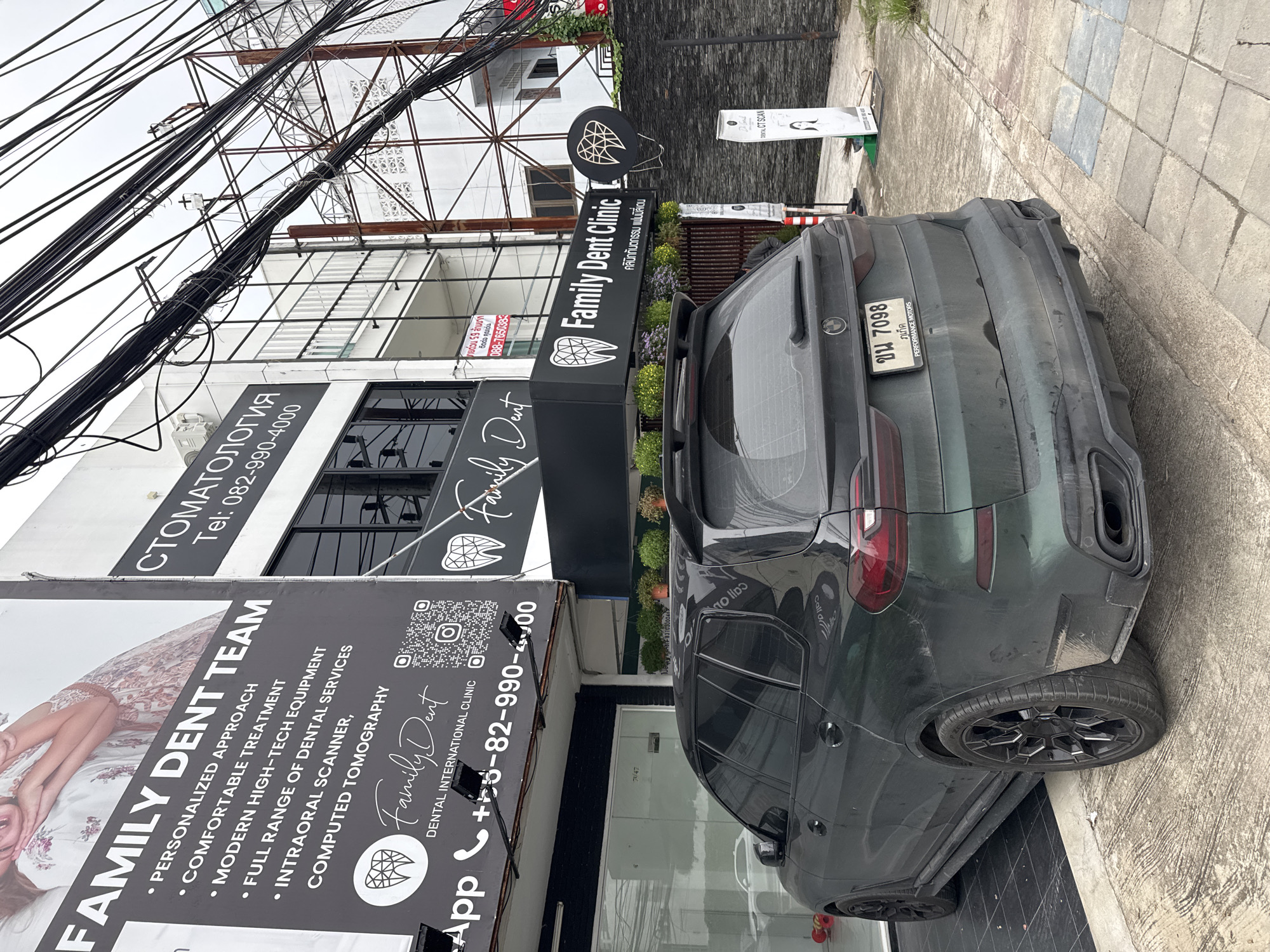 The image shows a dark green BMW SUV parked on a paved area in front of a building with multiple signs advertising a dental clinic. The building has a white facade with black lettering. One prominent sign reads "FAMILY DENT TEAM" with a list of services. Another sign displays "Family Dent Clinic" in English and Thai script. To the right, a vertical sign with an illustration of a woman and the text "DENTAL CT SCAN" is visible. Electrical wires hang overhead. The license plate on the SUV is visible and reads "ขม 7098". The scene appears to be on a street in Chalong, Thailand, indicated by the Thai script on the signs and license plate. No people are clearly visible in the image.
