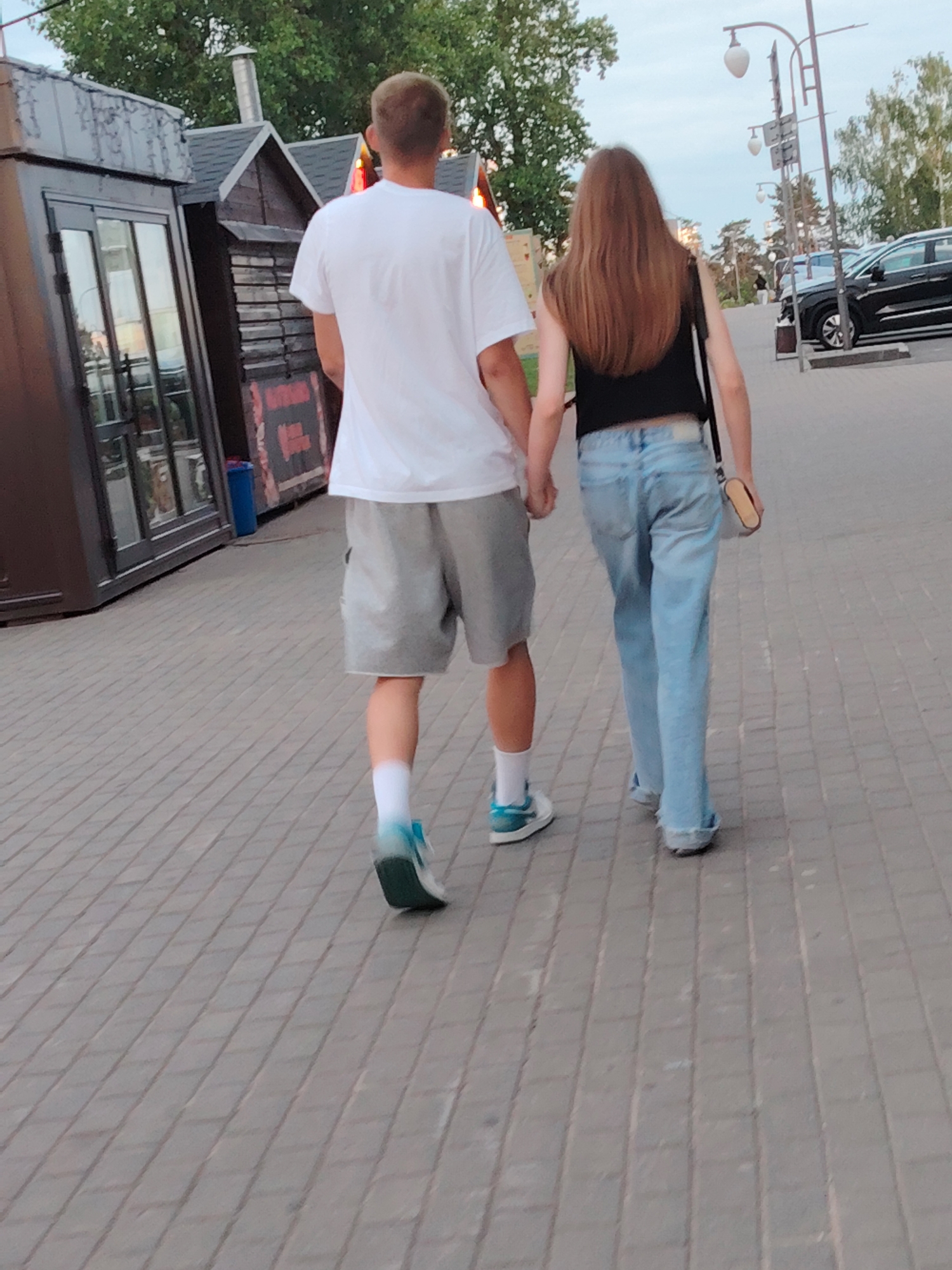 Two individuals, a male and a female, are observed from behind, walking hand-in-hand on a paved pathway in Бараўлянскі сельскі Савет, Belarus. The male, positioned on the left, has short, light brown hair and is wearing a white short-sleeved t-shirt, grey athletic shorts, white ankle socks, and light blue sneakers with white soles. The female, on the right, has long, straight, brown hair and is dressed in a black sleeveless crop top, light blue loose-fitting denim jeans with raw hems, and light-colored flat shoes partially obscured by the jeans. She carries a small, light-colored shoulder bag with a dark strap on her right side.
To the left of the individuals, there are two small commercial-style buildings: a darker, modern kiosk with a large glass door/window and a grey roof in the foreground, and a rustic wooden structure with a dark gable roof and a metal chimney behind it. A blue rectangular trash bin is situated between the buildings. A partially visible red and black sign is affixed to the wooden structure.
The pathway is composed of rectangular grey paving stones laid in a running bond pattern. To the right, a paved road or parking area is visible with several parked cars, including a dark SUV. Multiple dark streetlights with spherical white lanterns line the area. The background features numerous trees with green foliage under a light blue, overcast sky, suggesting a daytime or early evening setting.
