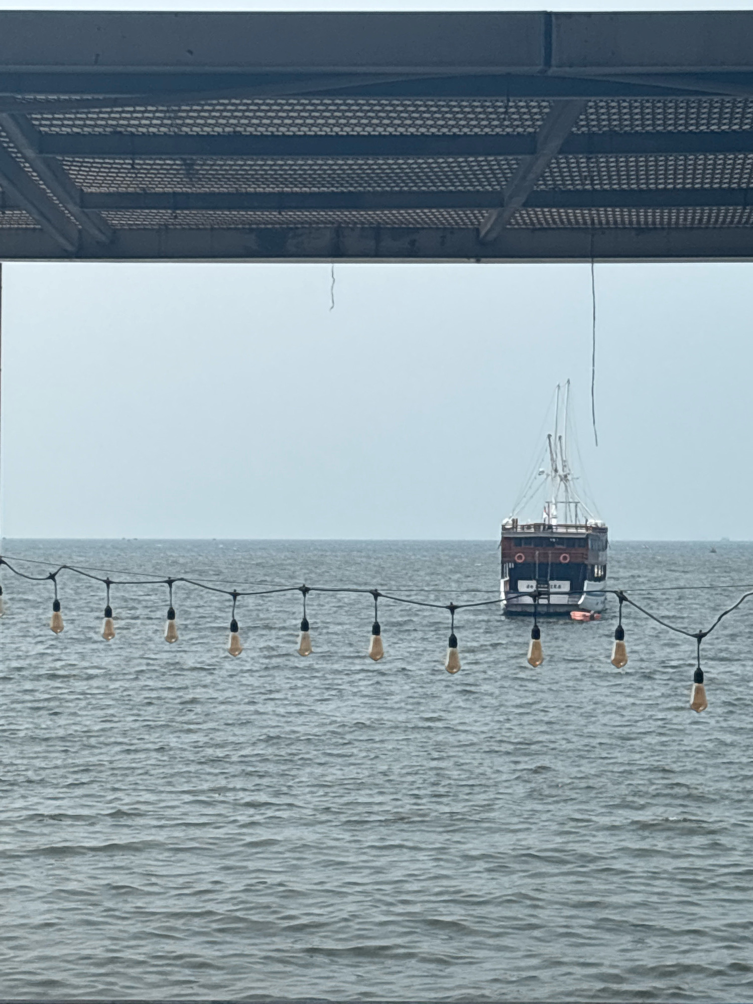 The image depicts a view of the sea from under an overhang, likely a pier or boardwalk, in North Jakarta, Indonesia. In the foreground, a string of decorative incandescent light bulbs hangs across the frame, illuminating the choppy grey water. In the distance, a two-masted schooner, possibly a tourist vessel, sails on the horizon under an overcast sky. The hull of the schooner appears to be made of dark wood and white siding, with circular portholes. A small orange life raft is visible near the stern. The water's surface is textured with ripples, suggesting a light breeze. The overhang above is a dark, metal structure with a perforated ceiling.