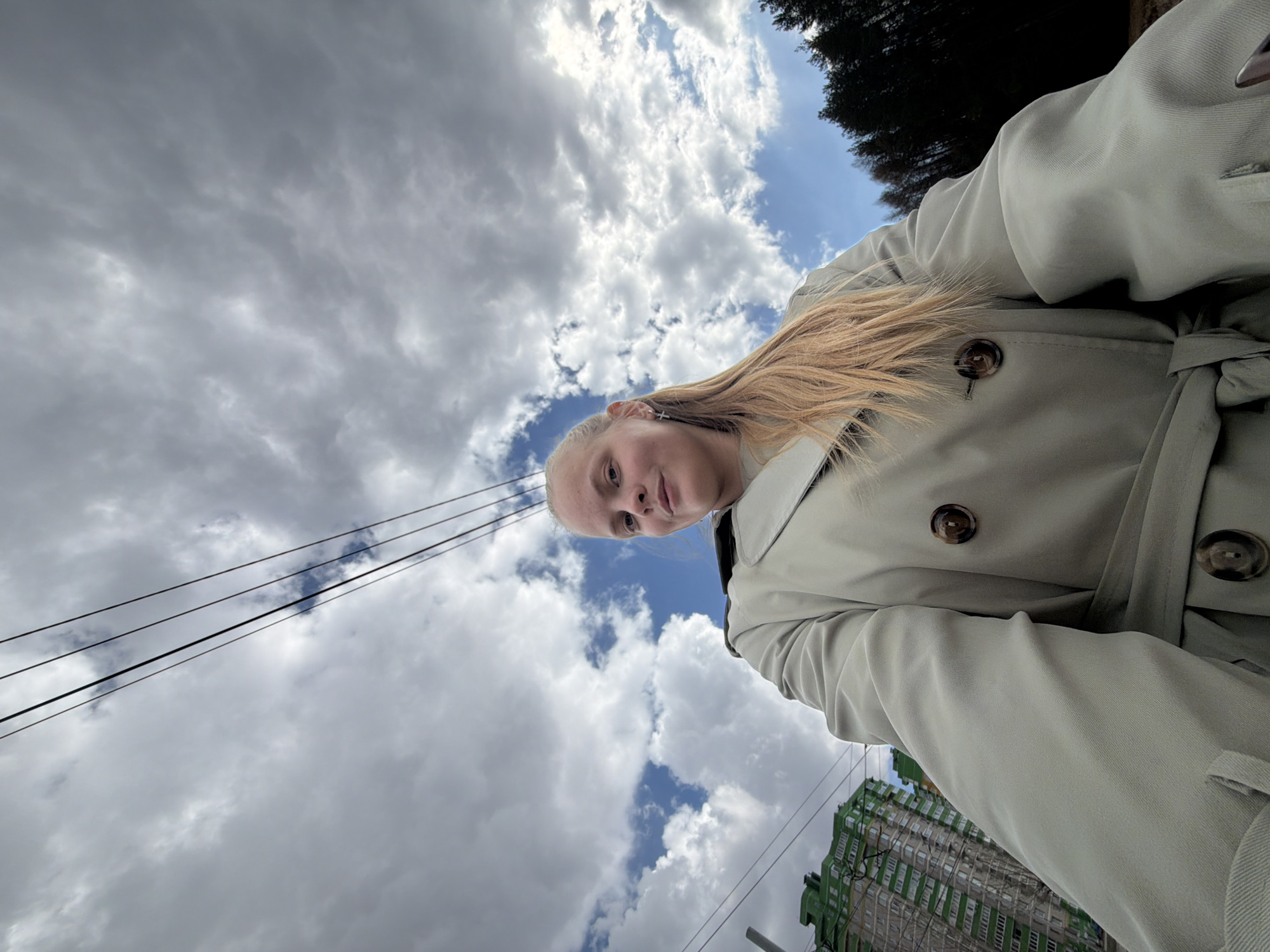 A young woman is depicted in a low-angle outdoor selfie, looking directly at the camera with a neutral expression. She has long, light blonde hair and fair skin, and is wearing a light-colored, possibly beige or pale green, trench coat with prominent dark buttons and a tied waist. A small cross-shaped earring is visible in her left ear.
The background is a dynamic sky, predominantly filled with large, bright white and grey clouds, interspersed with clear patches of blue sky, indicating it is daytime. On the upper right side, the dark green canopy of a tree is visible. Several parallel black utility wires stretch across the sky, particularly on the left side and towards the lower right. In the lower right portion of the image, behind the woman's arm and coat, a tall, multi-story building with distinctive green and grey/white architectural elements is partially visible. This building, along with the utility lines, provides urban context, likely in Izhevsk, Russia. The upward-looking perspective emphasizes the sky and creates a unique composition. There is no visible text in the image.