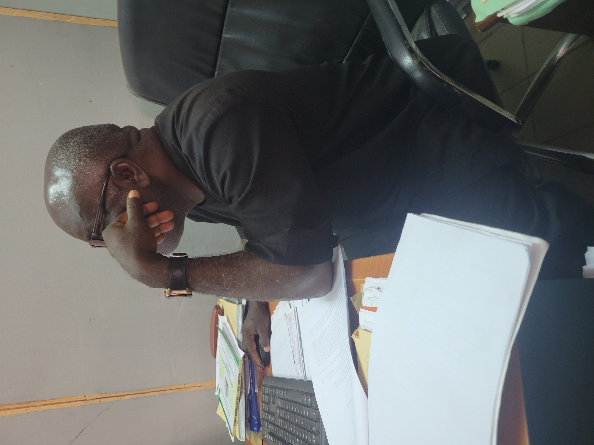 An indoor, eye-level shot in what appears to be an office or study, showing a dark-skinned man seated at a desk, in Abuja, Nigeria. The man, possibly bald or with very short hair, wears glasses and a black short-sleeved top. He is positioned facing right, with his right hand resting on his chin or mouth, suggesting contemplation, weariness, or deep thought. A black watch with a dark strap is visible on his left wrist.
The man sits on a black office chair with armrests. In front of him, a wooden desk is cluttered with various documents, papers, and folders. A black computer keyboard and a mouse are partially visible on the desk. Among the visible documents, one green-covered book clearly displays the title "PUBLIC SERVICE RULES," and another document or folder beneath it shows "2001 EDITION." The general setting indicates an active workspace. The background consists of a plain light grey wall. The lighting suggests an indoor environment, likely office lighting.