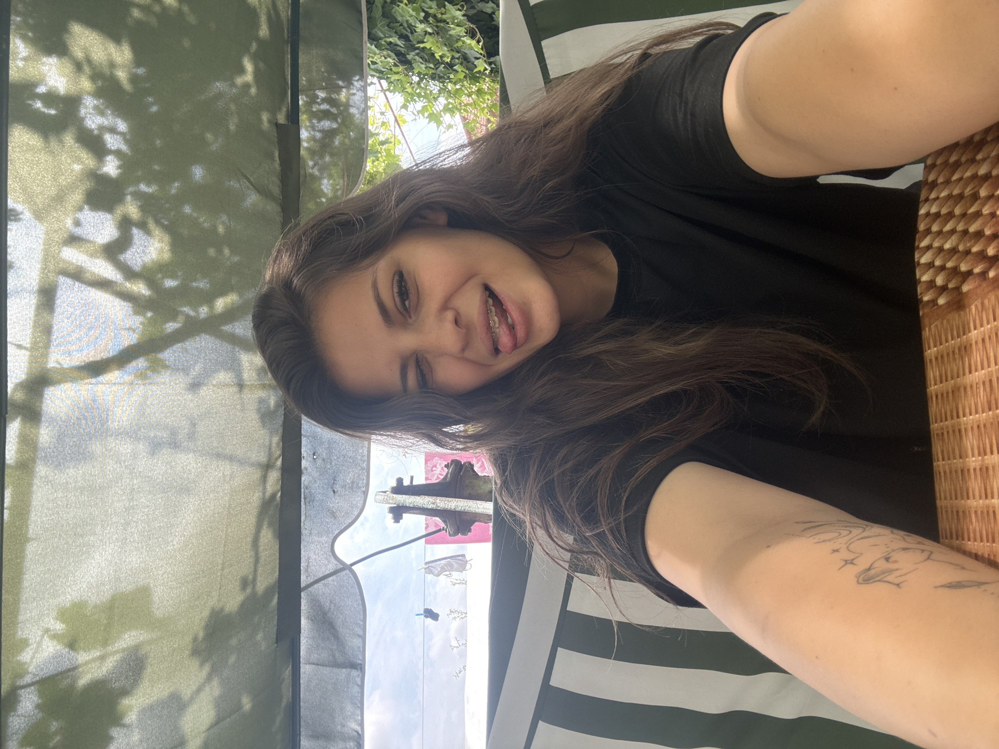 A young woman with long brown hair and braces is sitting under a canvas canopy, possibly a swing or gazebo, outdoors. She is wearing a black t-shirt and sticking her tongue out while winking. Her left arm, visible in the foreground, has a tattoo. In the background, there is a clothesline with laundry hanging, a metal pole, and lush green ivy. The scene suggests a casual, relaxed setting, potentially in a backyard or garden. The location is the village of Govedare, Bulgaria.