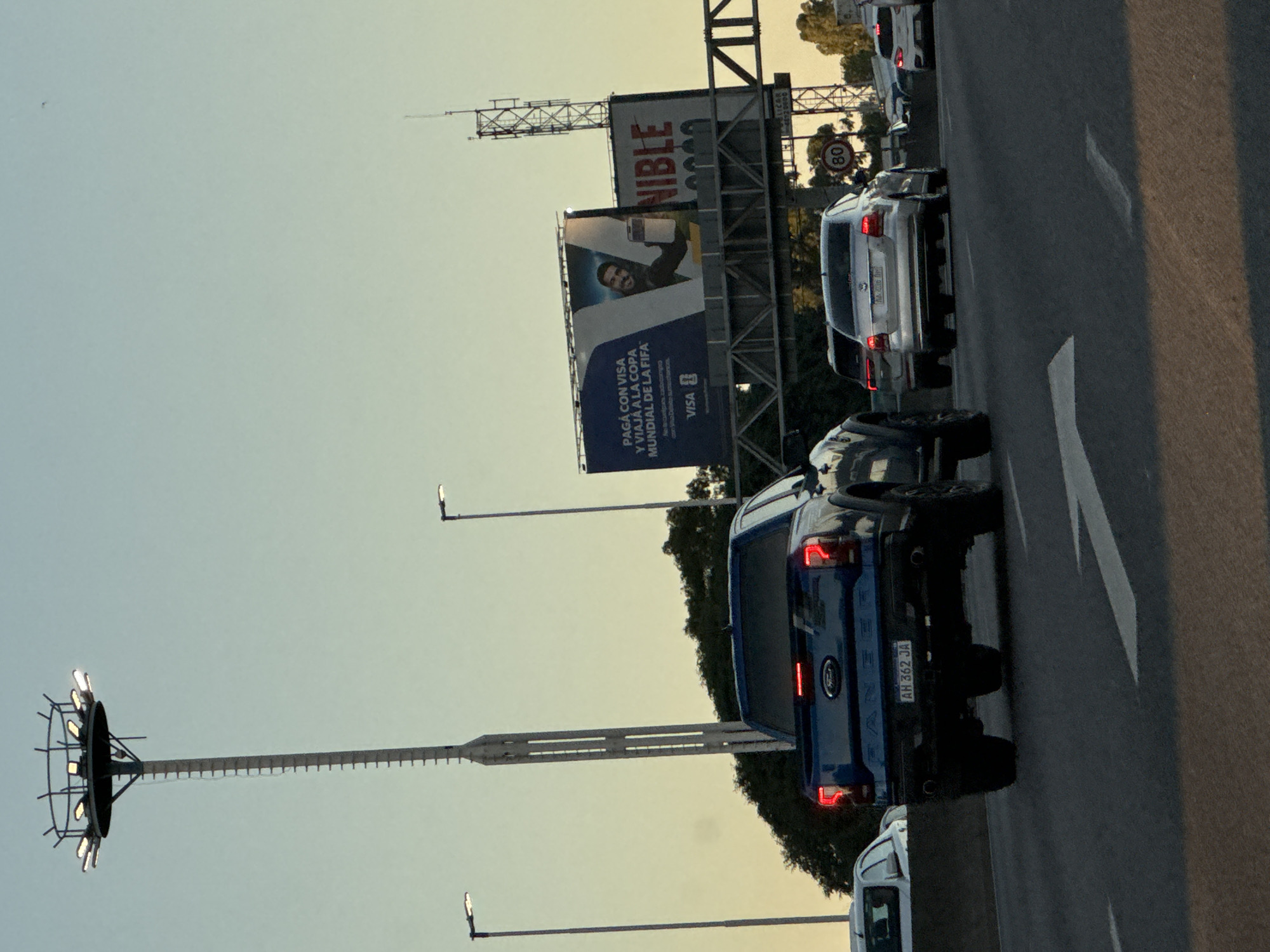 The image captures a congested traffic scene on a road in Buenos Aires, Argentina. The camera angle is oriented at a rightward tilt. The sky is washed with a soft, faded yellow-orange sunset. The road is lined with a queue of vehicles, including a blue Ford Ranger visible in the foreground, a white van in the lower part of the frame, and multiple cars ahead. The license plate "AH 362 JA" is clearly visible on the Ford Ranger.
Dominating the background is a large billboard advertising VISA for the FIFA World Cup with the tagline "PAGA CON VISA Y VIAJA A LA COPA MUNDIAL DE LA FIFA" and the brand name "NIBLE" above it. A circular road sign indicates a speed limit of 80 km/h. A tall lighting structure, equipped with a circular array of lights, stands along the roadside. Utility poles are also visible.
